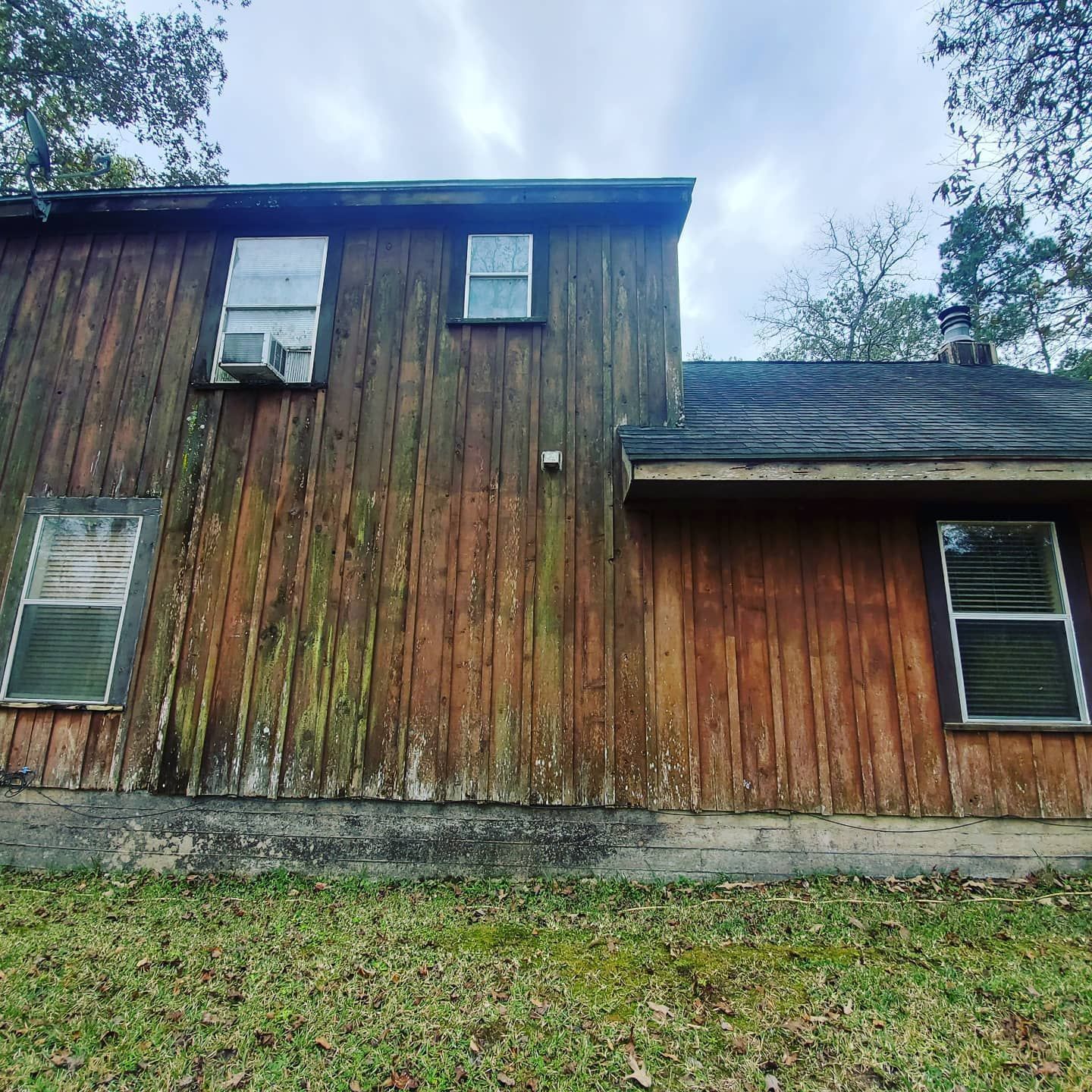 Weathered wooden house exterior with two stories, small windows, and a grassy yard under cloudy sky