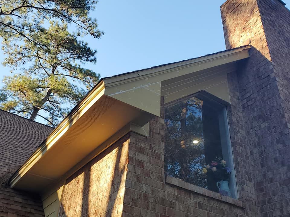 Brick house exterior with a large window, sloped roof, and tall pine trees against a blue sky