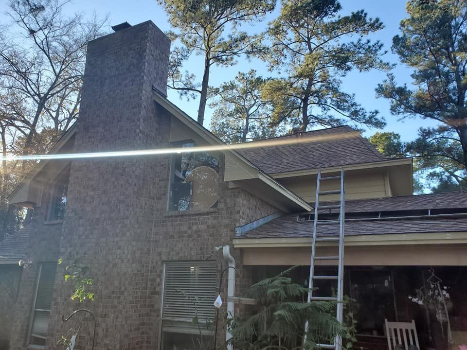 Brick house with chimney, covered porch, and ladders leaning against the roof under tall trees