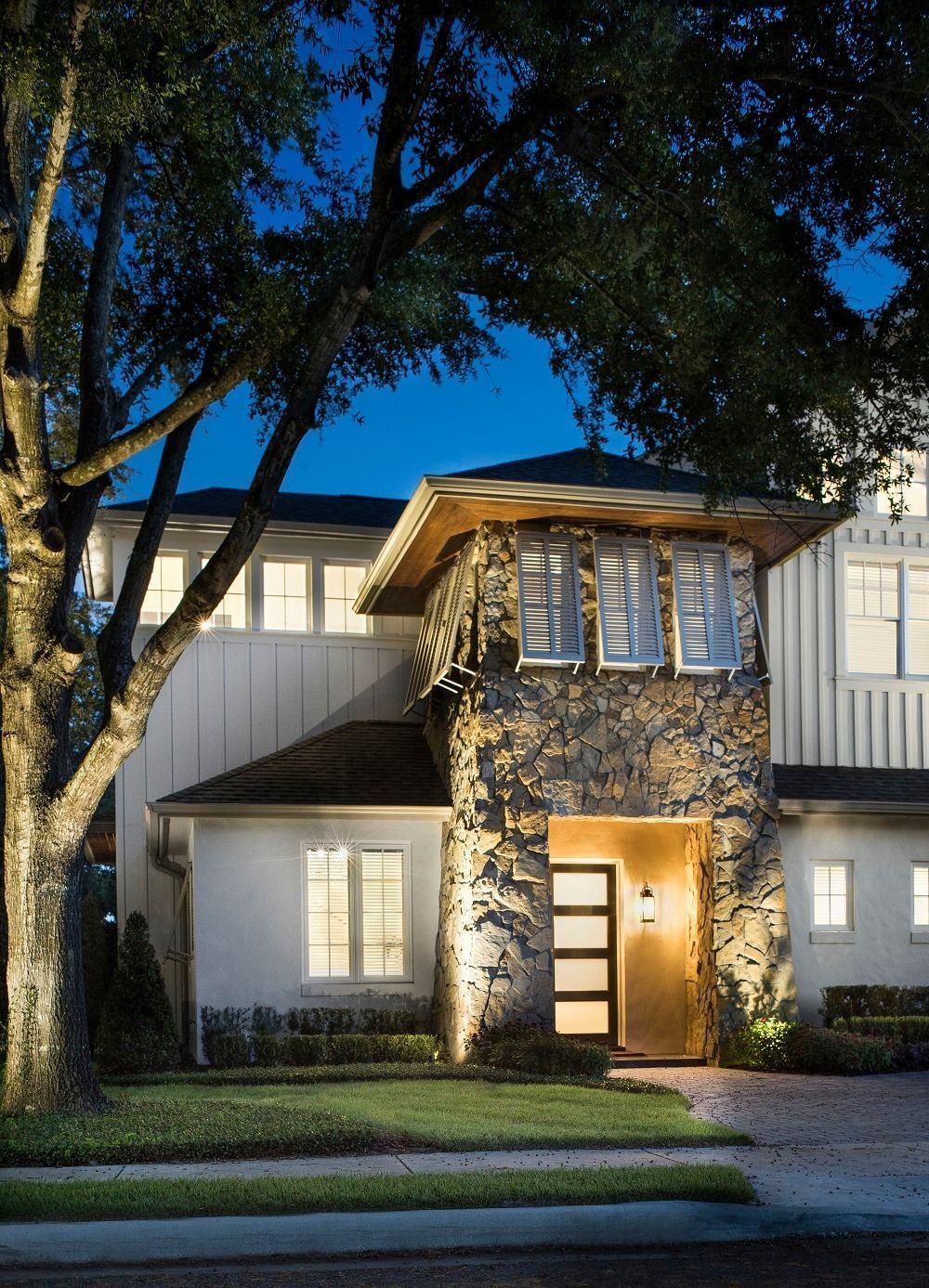 Two-story house at dusk with lit front entrance and stone column, framed by large trees