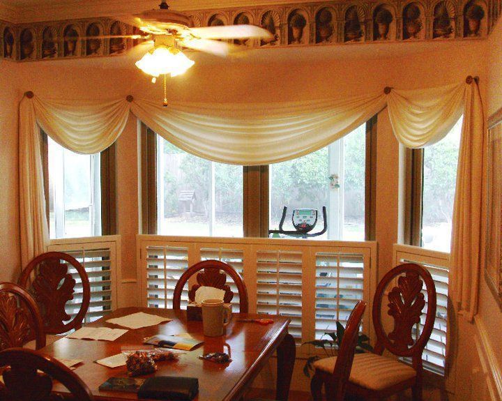 Warm dining room with large bay windows, cream curtains, wooden chairs, and a sunlit table.