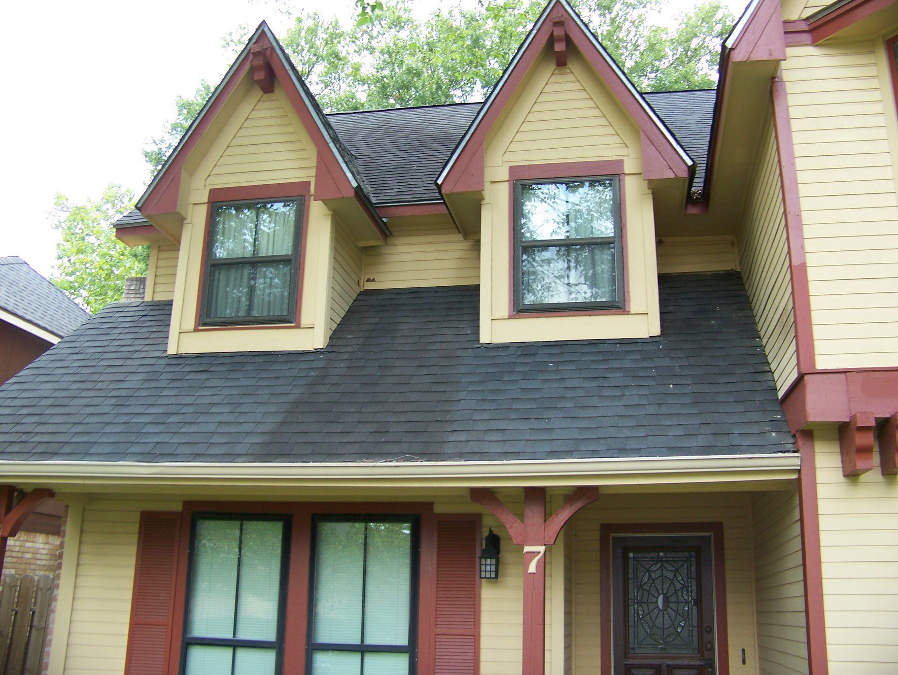 Yellow house with dark roof and two dormer windows under a leafy tree.