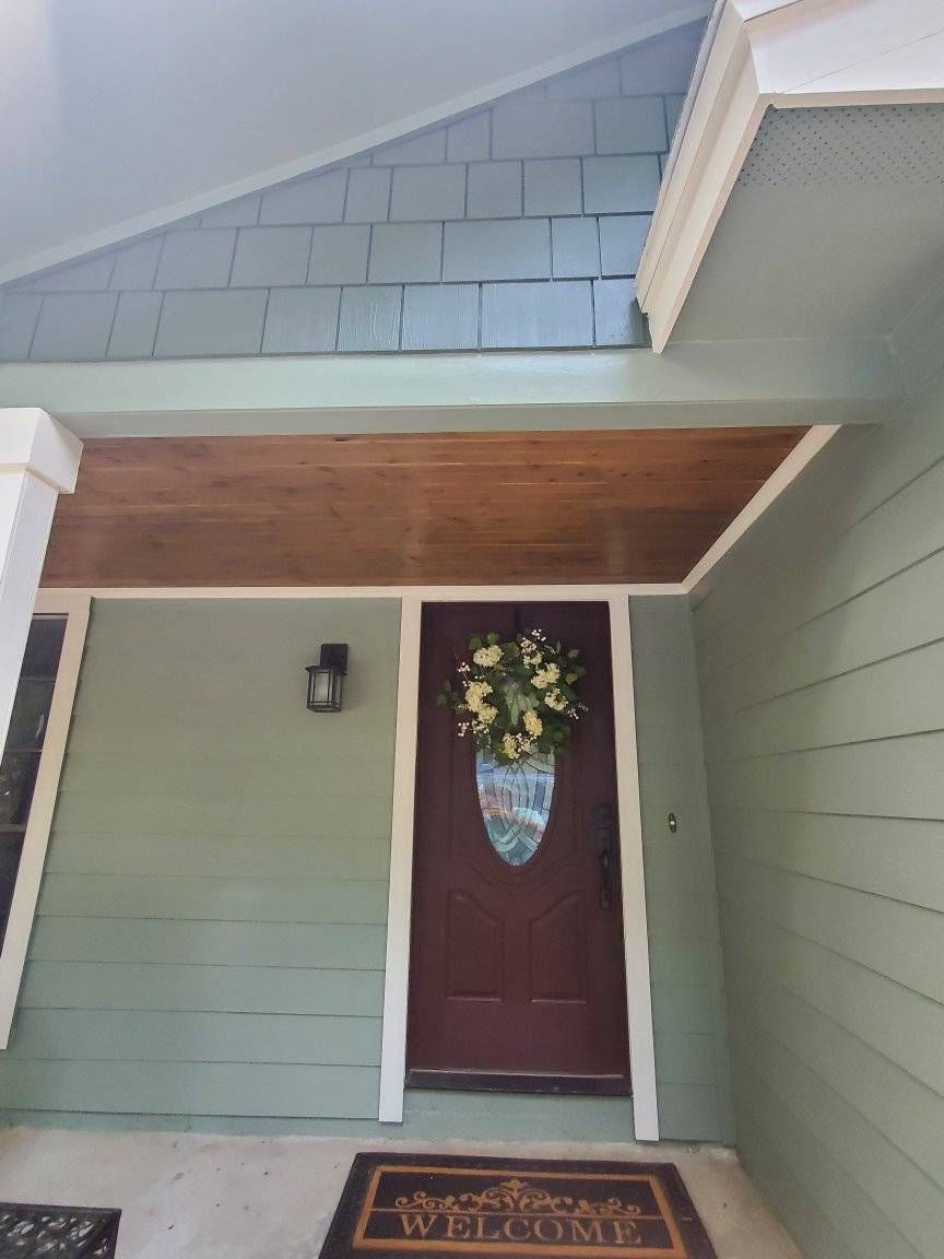 Front porch with green siding, maroon front door, wreath, and doormat under a covered roof