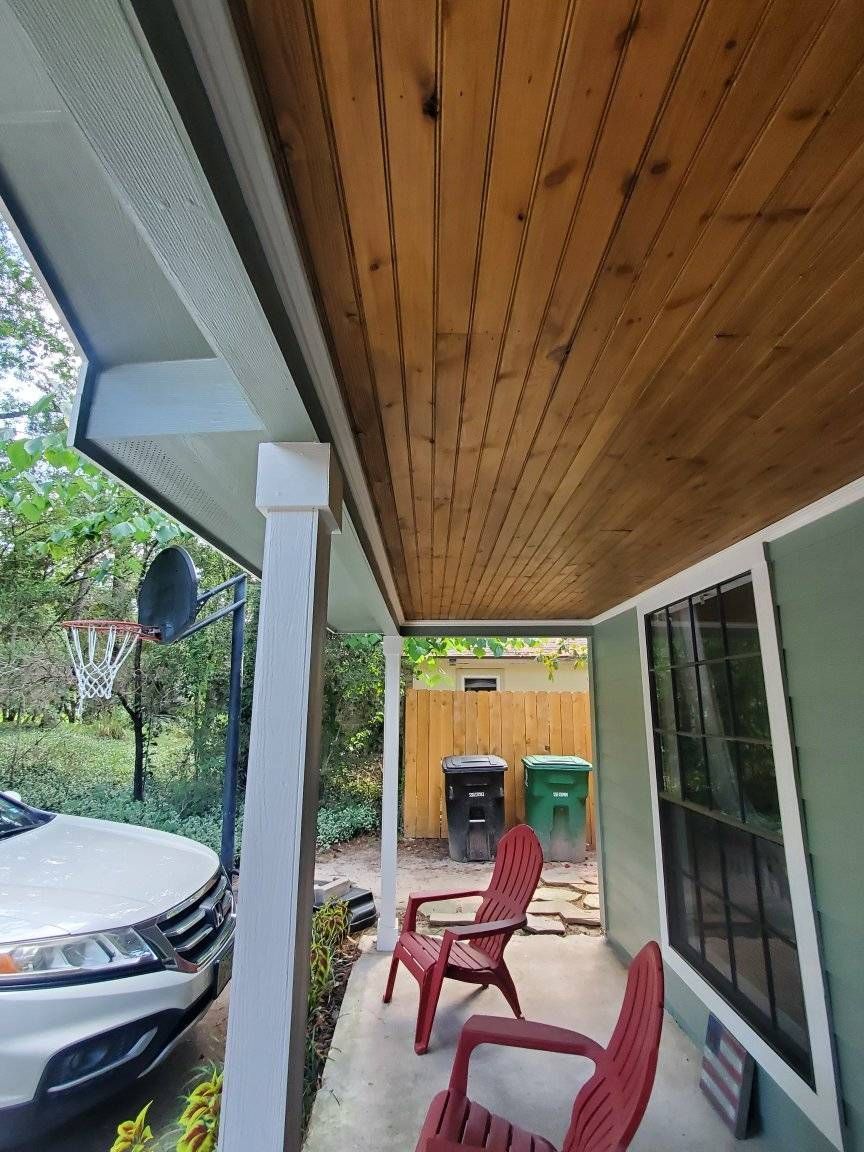 Covered porch with red chairs, car parked beside it, and trash bins near a wooden fence.