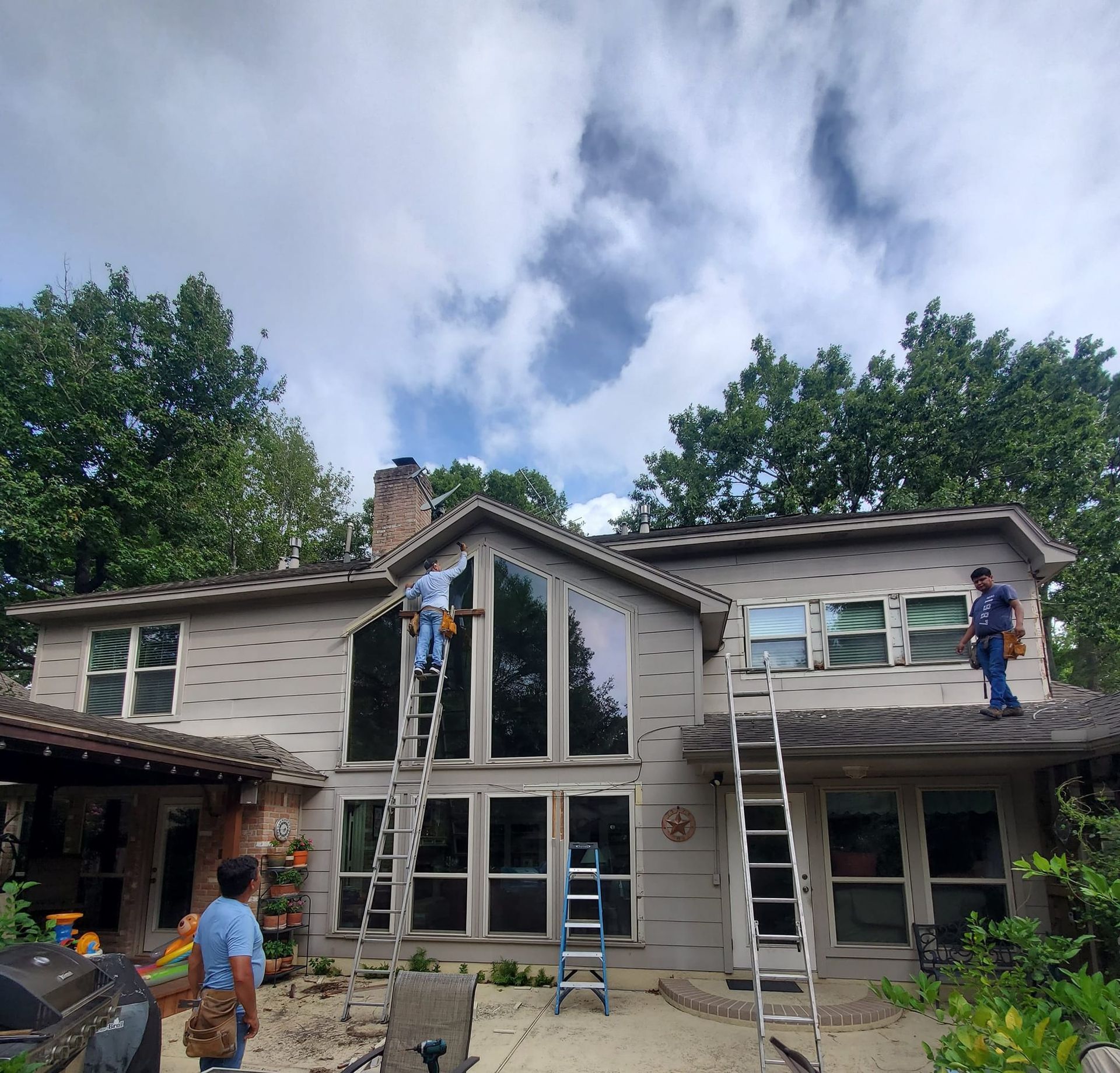Workers on ladders repairing a two-story house with large windows and a stone chimney