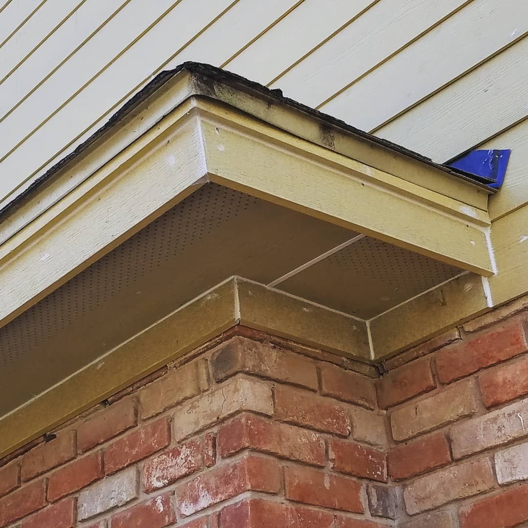 Close-up of a house roof corner with yellow siding, brick wall, and a blue gutter bracket
