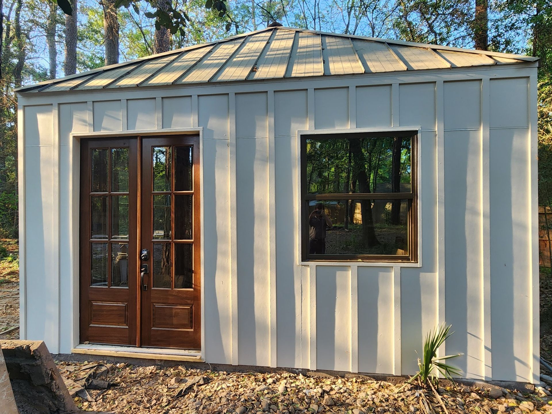 Small cream shed with a metal roof, wood double doors, and a window among trees.