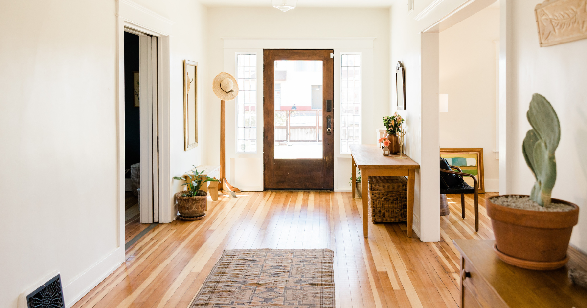 Bright hallway with hardwood floors, rug, wooden table, potted plants, and a front door at the end