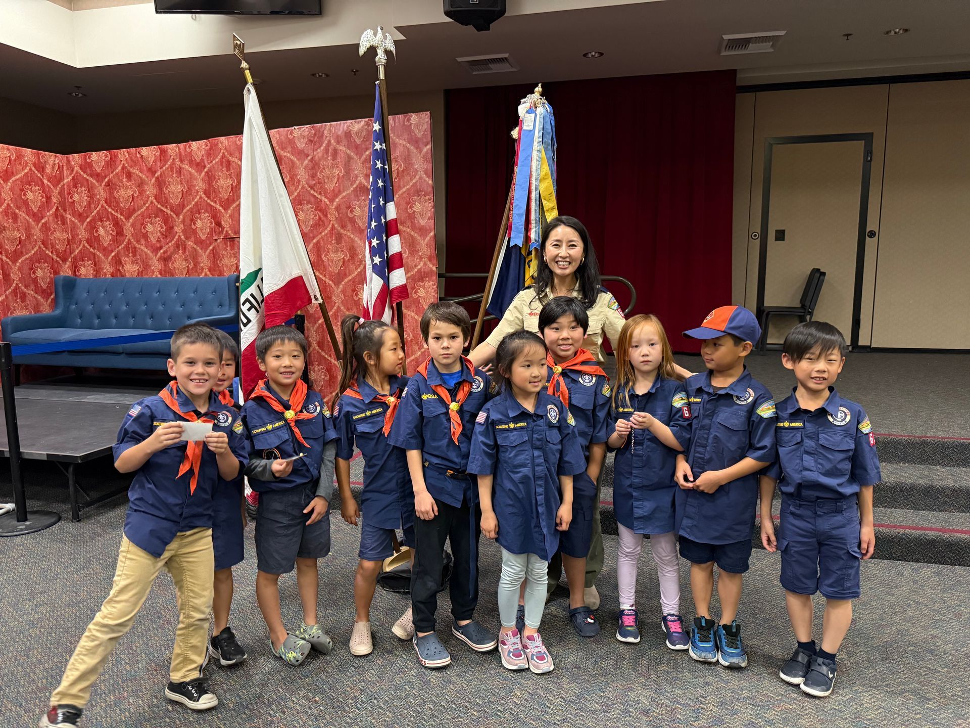 Child scouts posing on a stage with flags, smiling in navy uniforms.