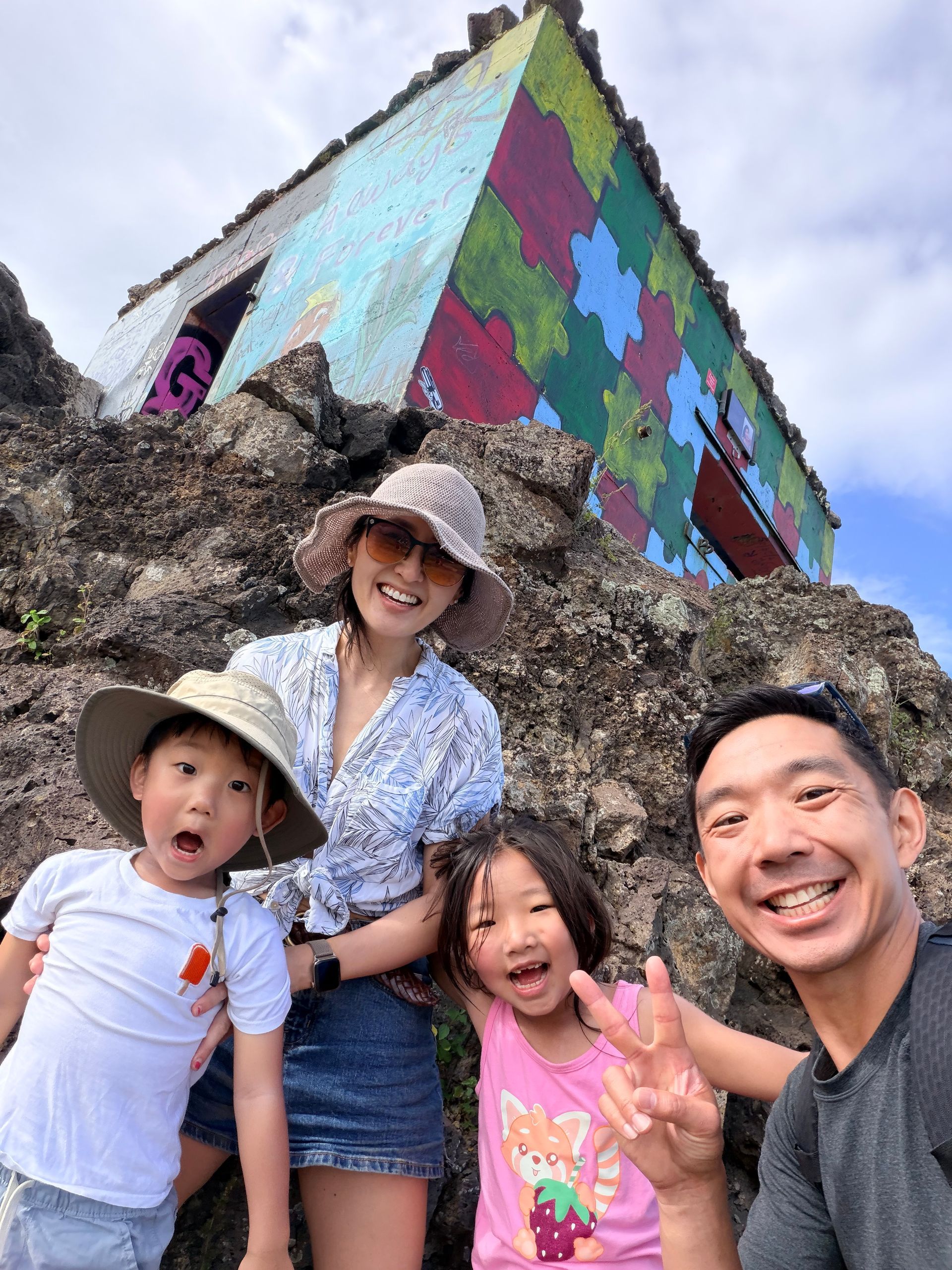 Family posing in front of a colorful geometric mural on a rocky hillside, smiling and making peace signs