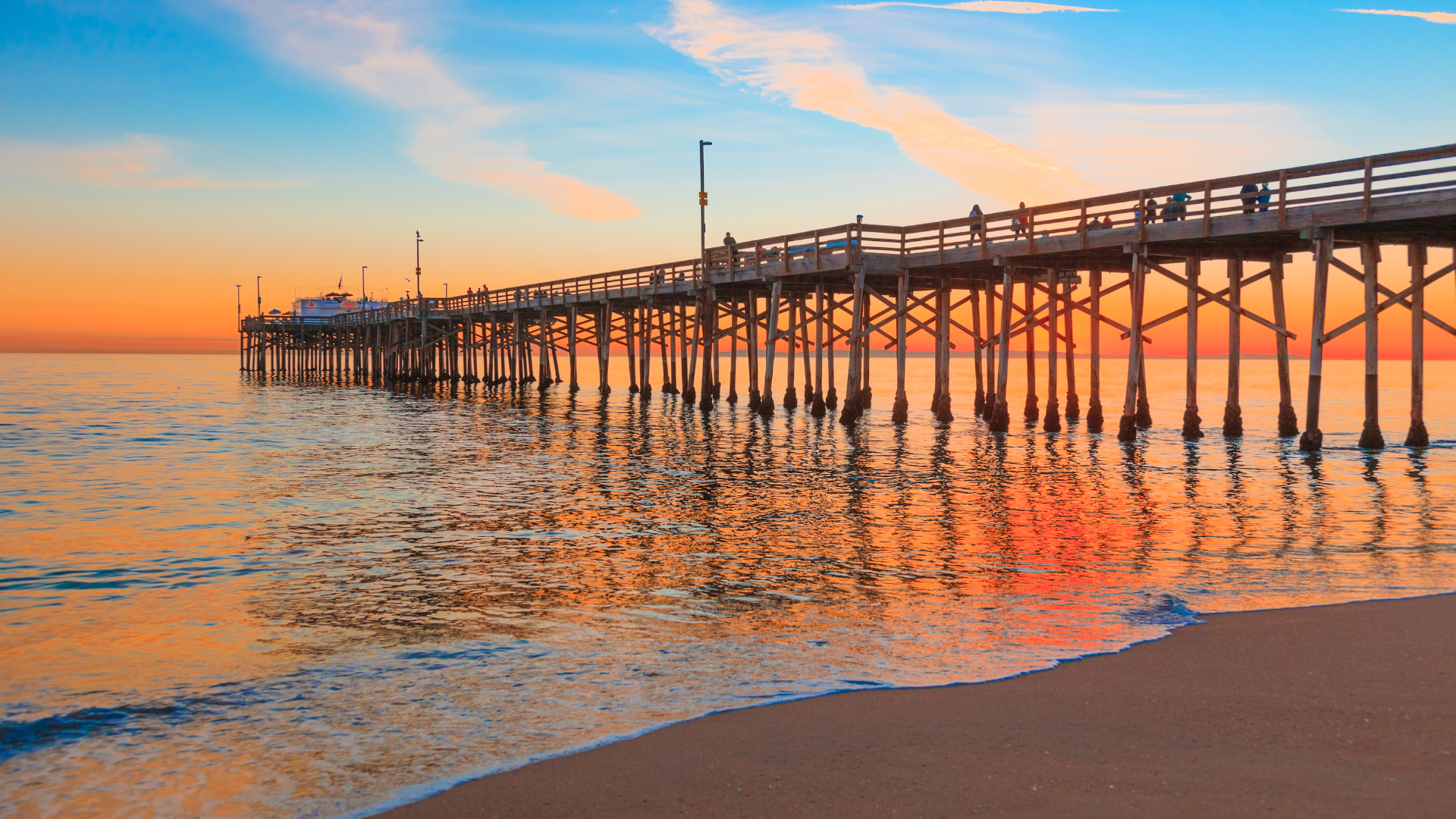 Sunset over a wooden pier stretching across calm ocean waves, with orange and blue reflections on the water