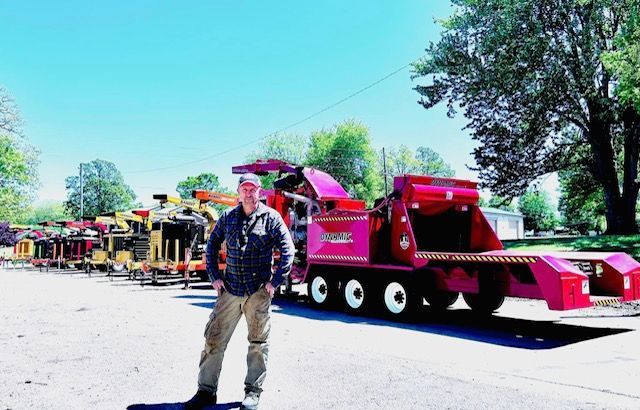 Man standing by a bright red amusement train on a sunny street, with colorful cars lined up behind him