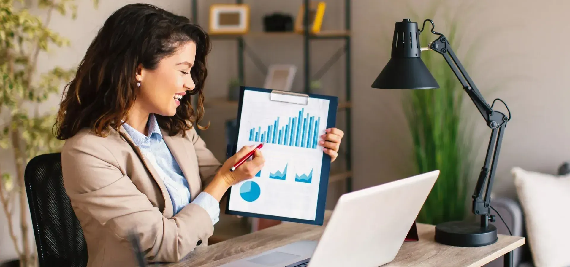 A woman is sitting at a desk with a laptop and holding a clipboard with a graph on it.