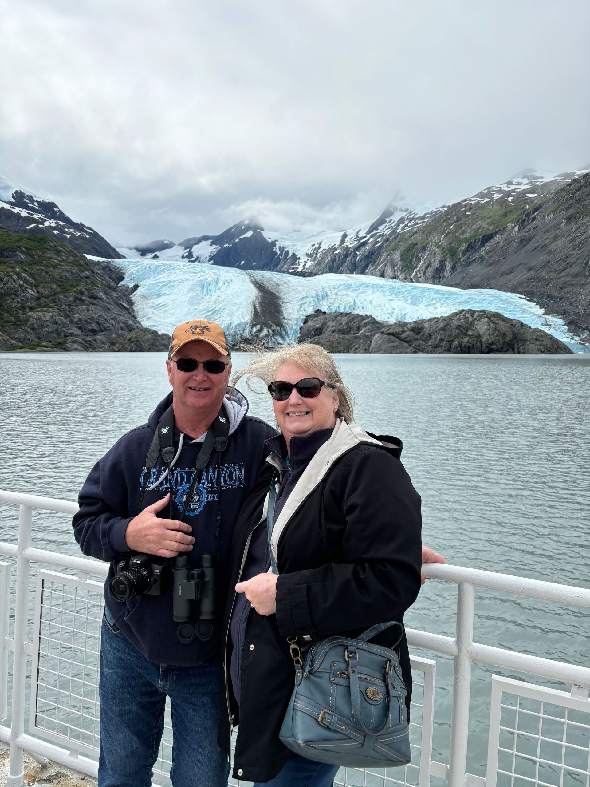 A couple poses on a boat deck with a large, icy blue glacier and mountain range in the background.