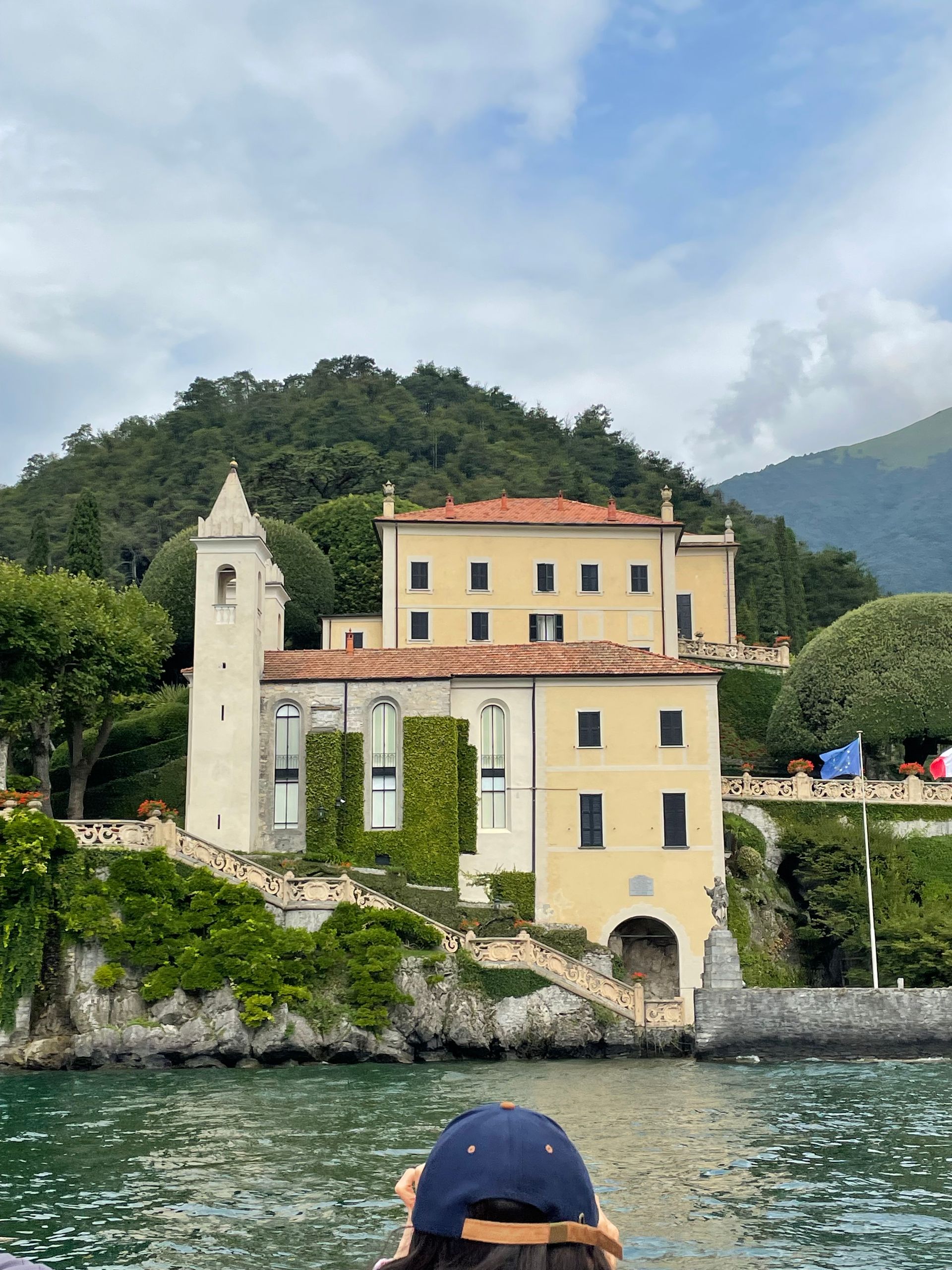 A historic yellow villa with a stone bell tower sits on a rocky shoreline overlooking a lake with a person in the foreground.