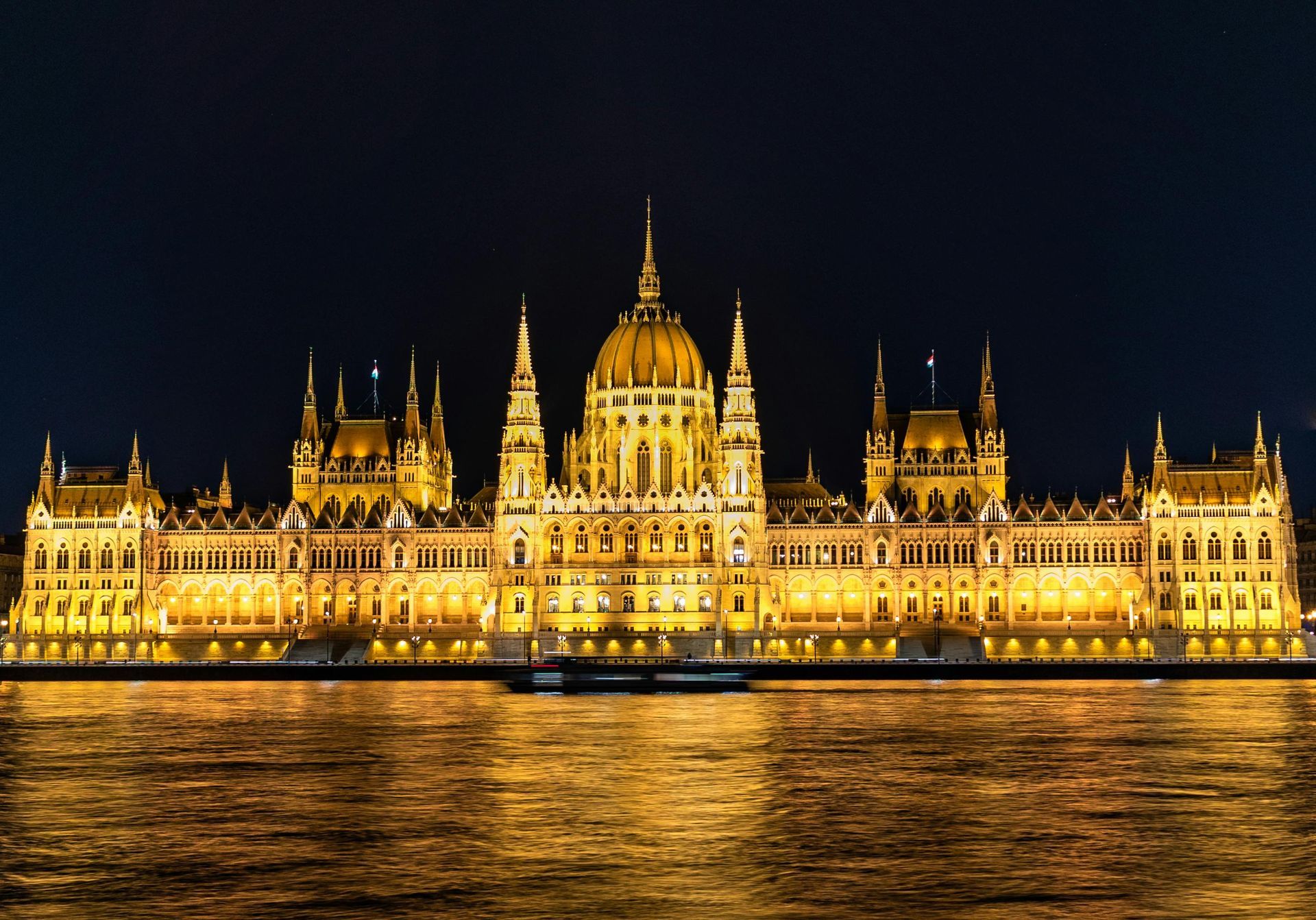 The illuminated Hungarian Parliament Building at night, viewed from across the Danube River with reflections on water.