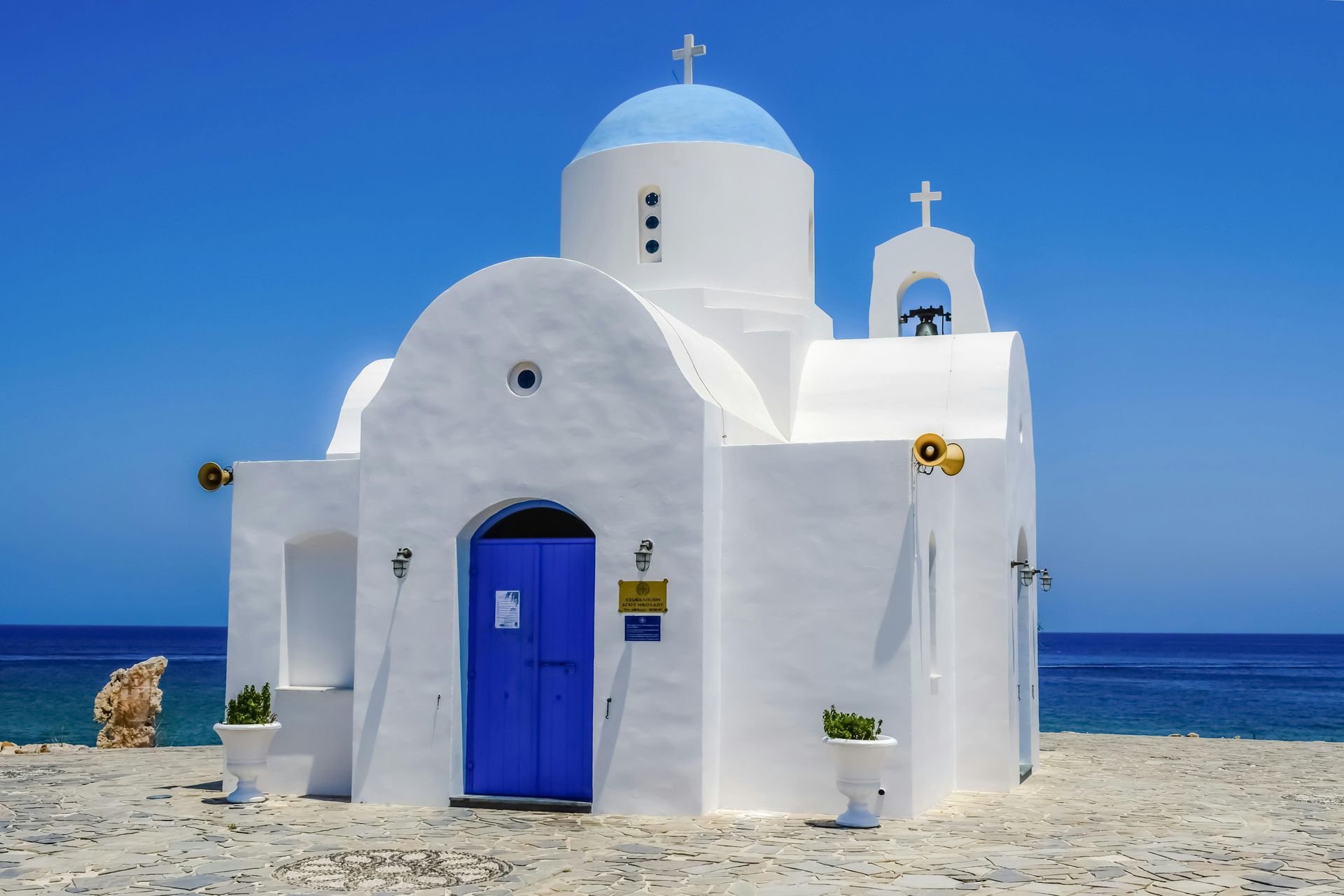 A white church with a blue dome and matching blue door, set against a rocky beach and the deep blue sea under a clear sky.