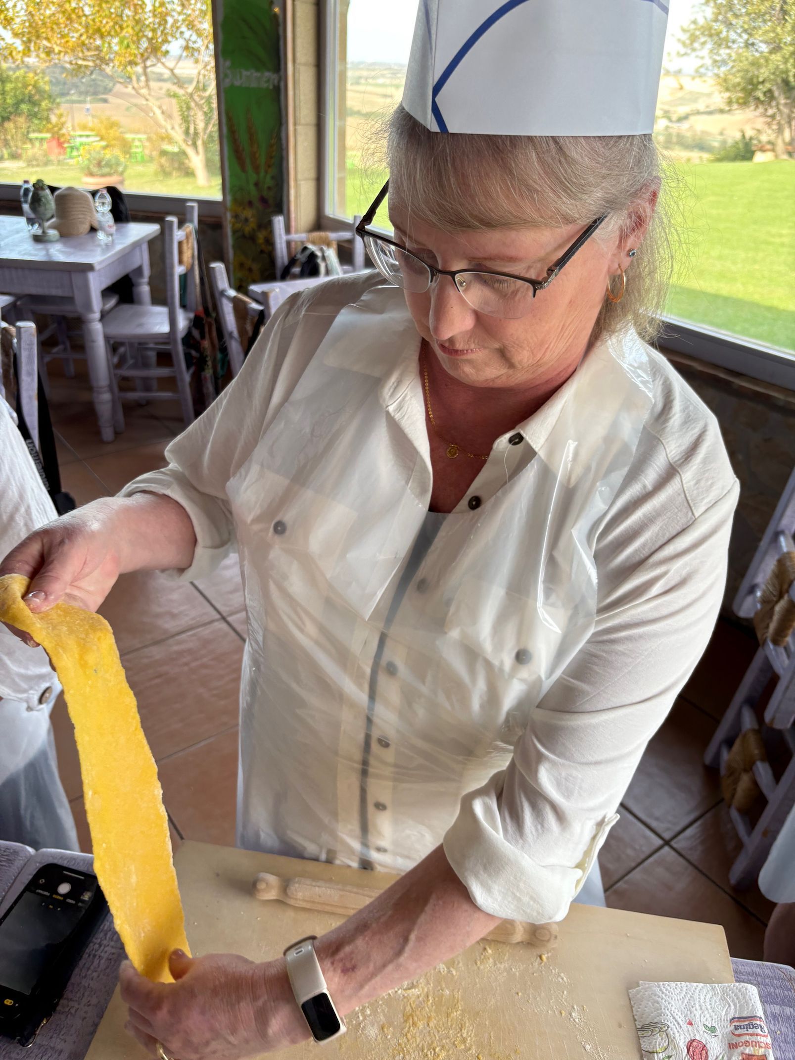 A person in a white chef’s uniform and hat holds a long strip of yellow pasta dough over a wooden surface.