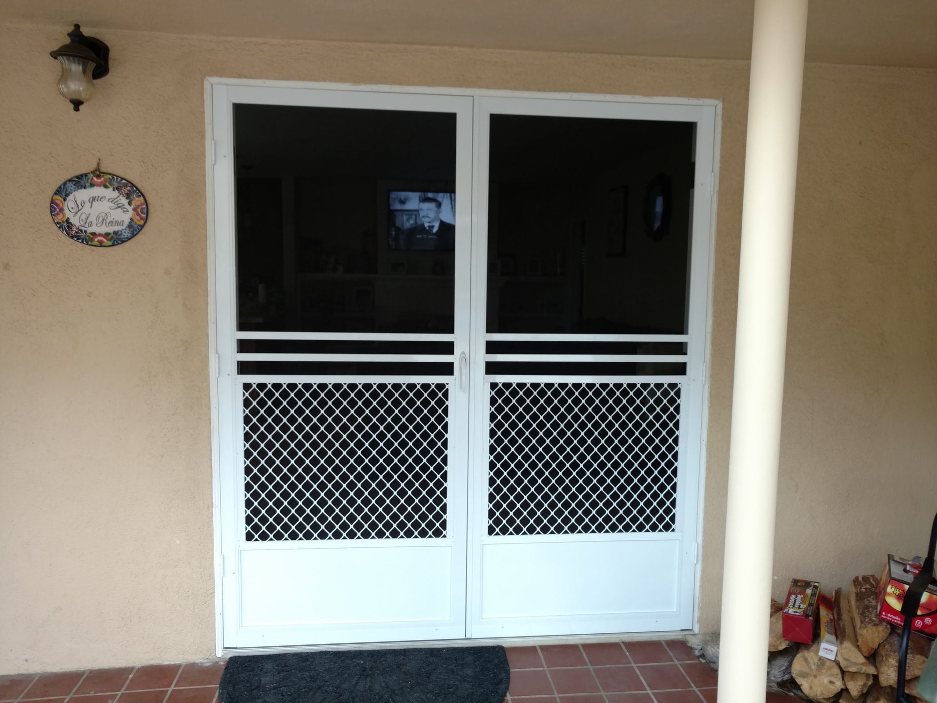Front porch with beige wall, white double screen door, wall lamp, decorative plaque, and potted flowers.