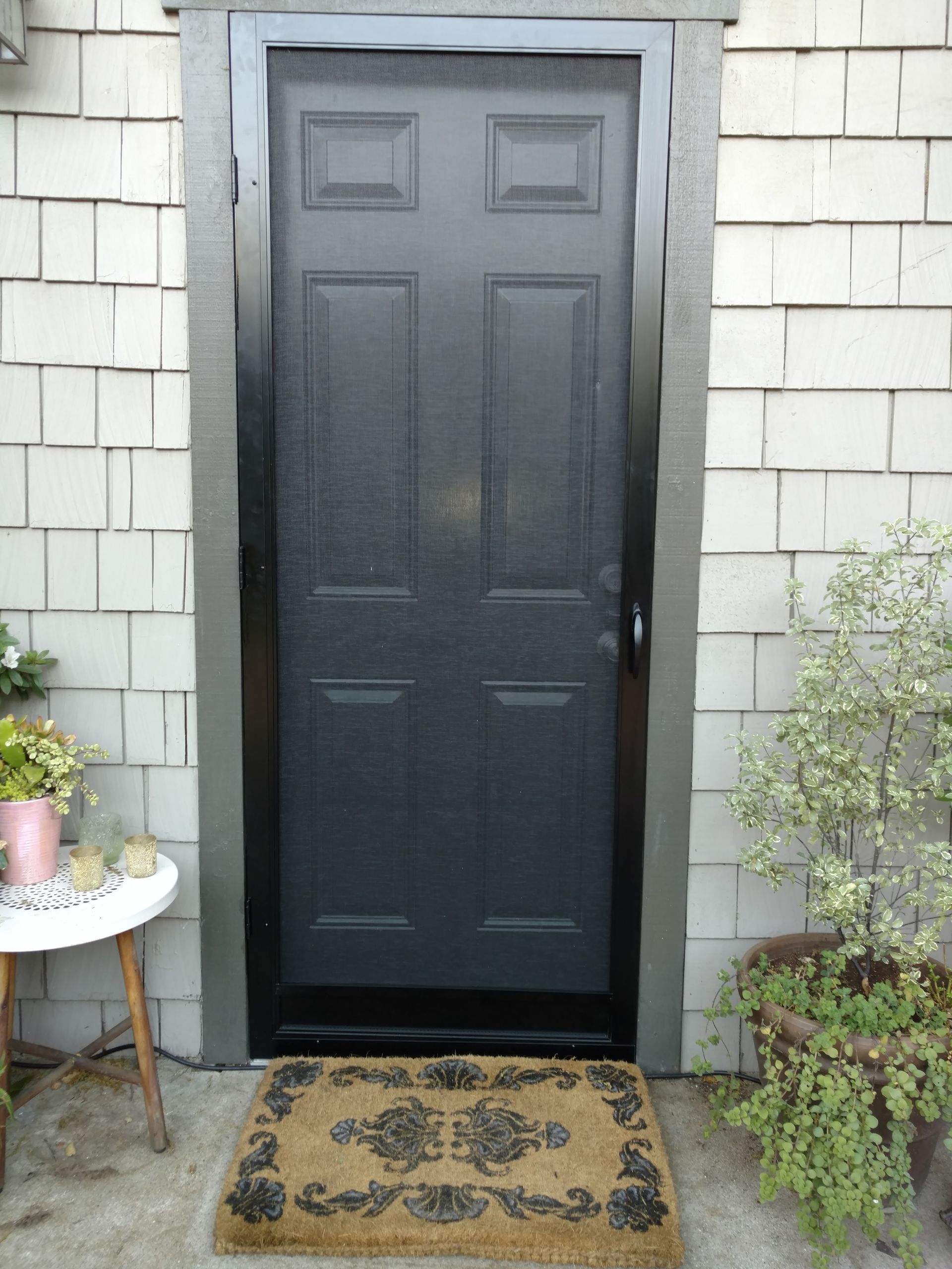 Black front door with a doormat, flanked by potted plants against gray shingle siding.