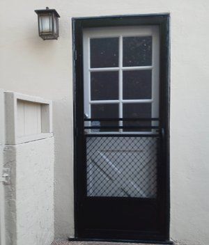 Black-framed door with a window and wire mesh panel set in a white wall, with a lantern beside it.