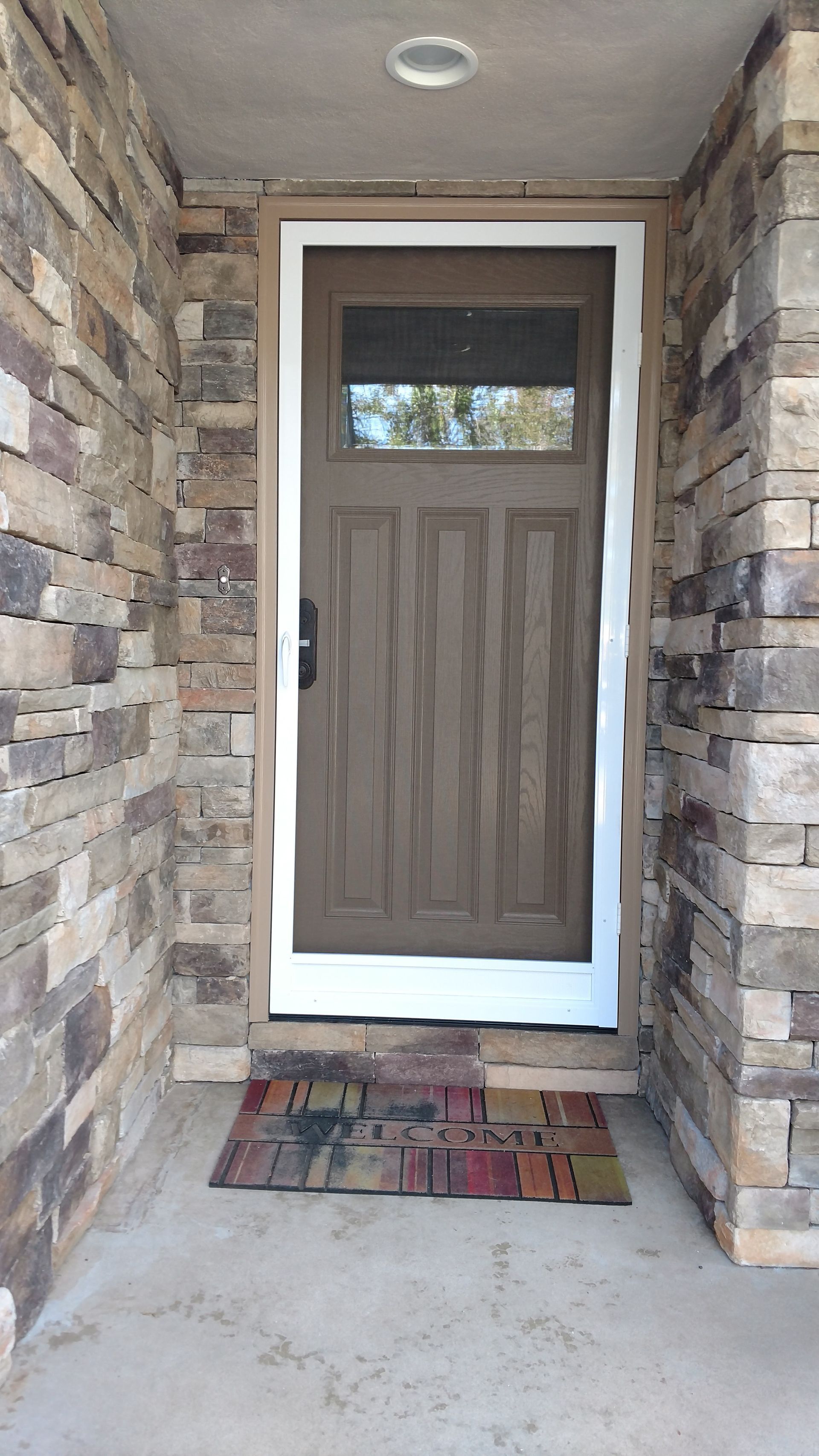 Front door with white trim and tan panels, centered in a stone entryway with a small doormat.