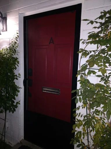 Red front door with a mailbox slot, framed by white siding and plants on both sides.