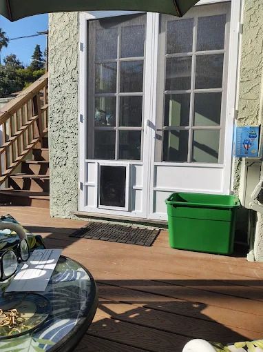 Deck with white French doors, green storage bin, and umbrella-shaded table in sunlight