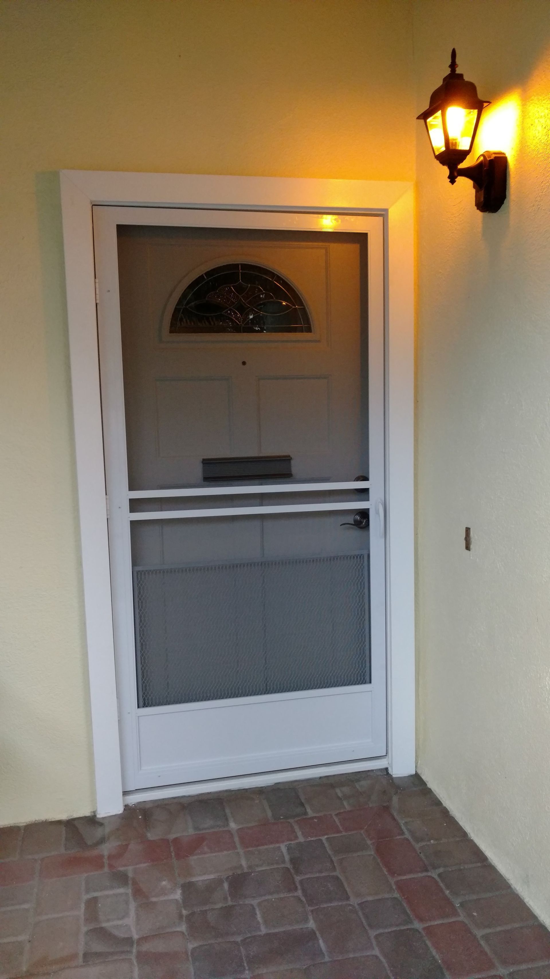 White screened front door with arched window beside a lit wall lantern on a porch with brick floor