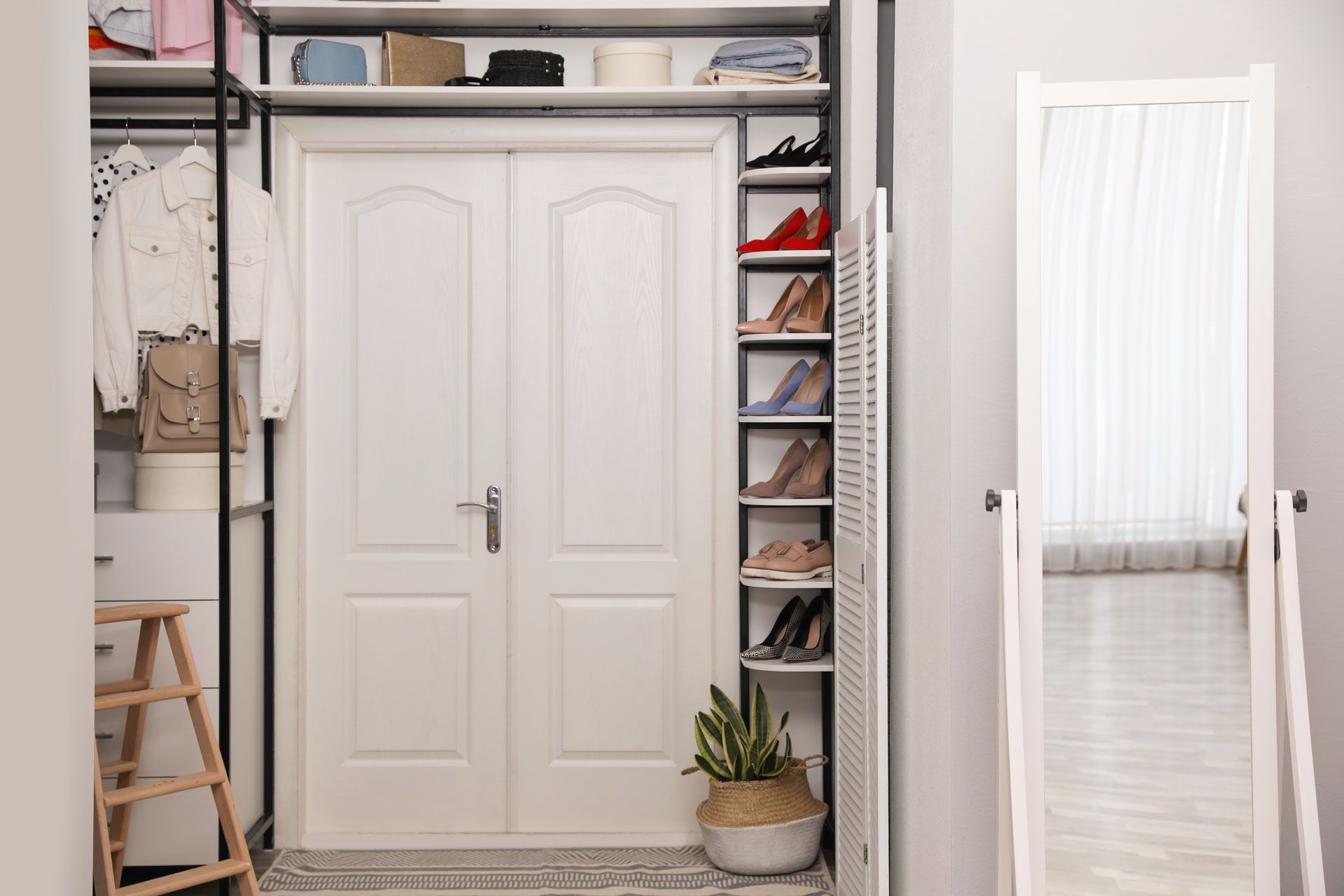 White closet with open shelves, hanging clothes, shoes, a stool, and a potted plant in a bright hallway