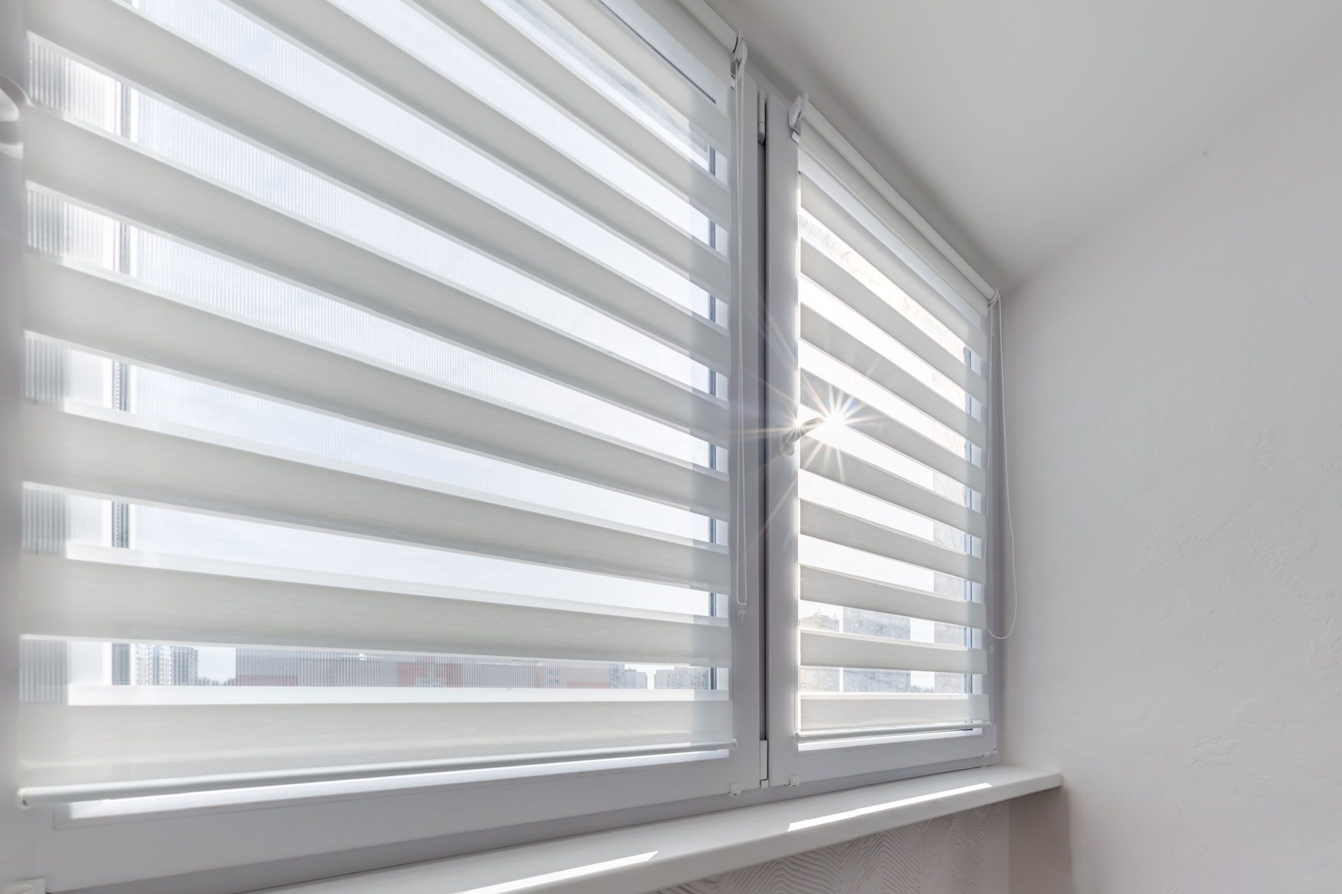 White zebra blinds on two windows in a bright room, with a white sill and wall.