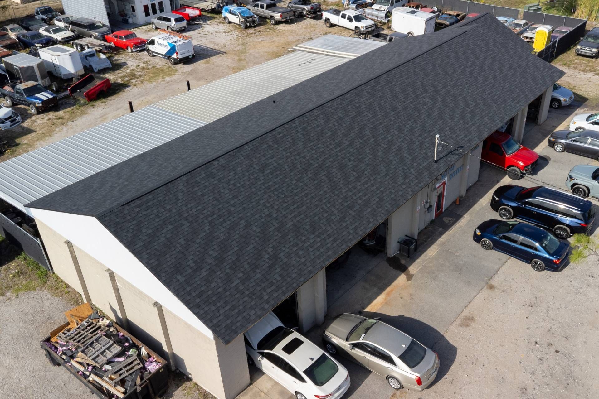 Aerial view of a long automotive shop building with a dark shingled roof, surrounded by cars and a storage lot.