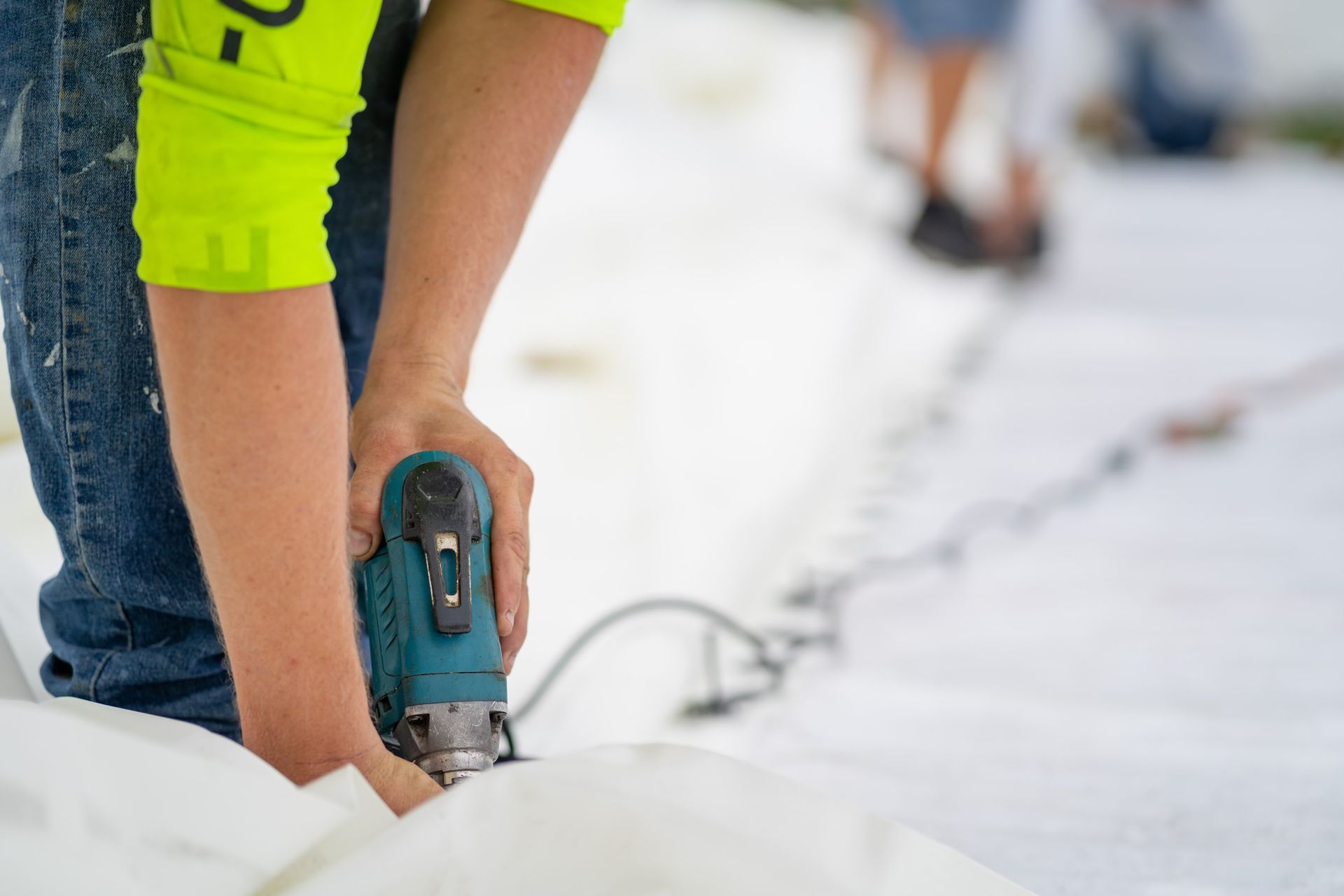 A person in a neon yellow sleeve uses a teal power tool to secure a white tarp to the ground.