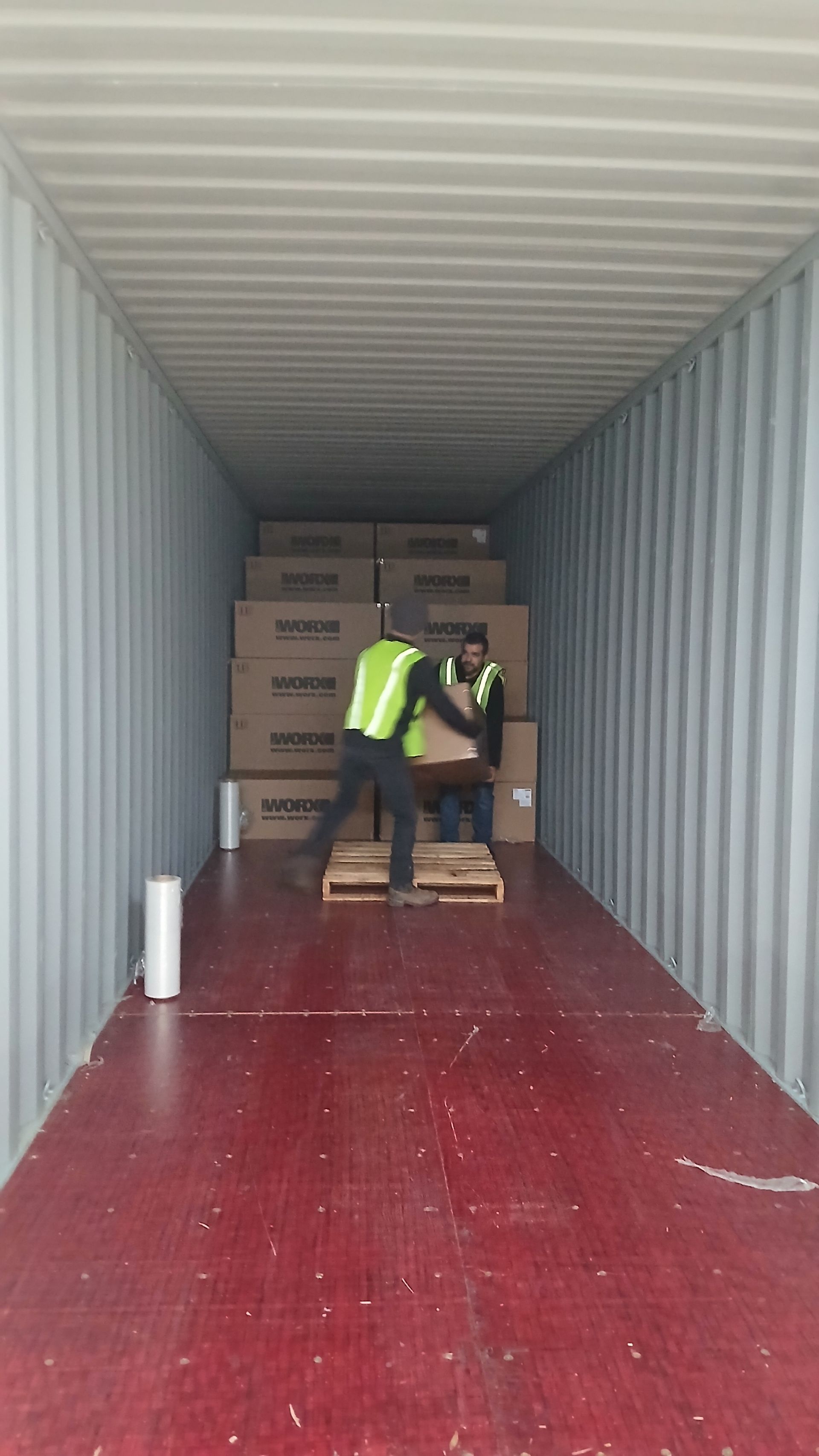 Two workers loading boxes onto a pallet inside a shipping container. One wears a reflective vest.