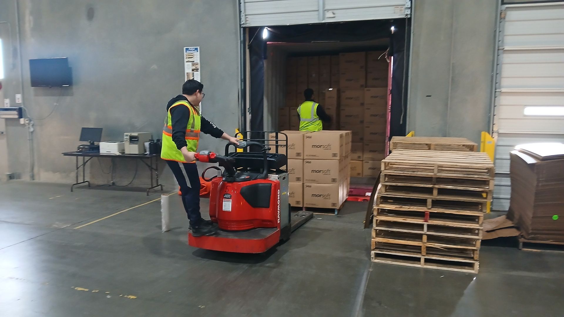 Person operating a red pallet jack inside a loading dock, moving boxes. Another person unloads boxes in the dock.