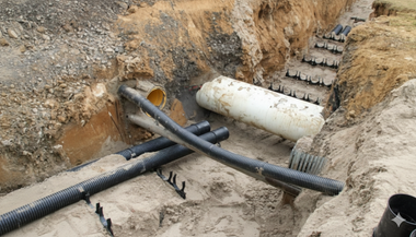 Trench dug for drainage with corrugated black pipes, yellow pipe, and a white cylinder on sand.