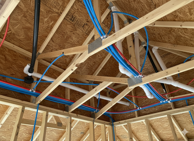 Blue and red pipes, and black drain pipes run through wooden rafters of a building's unfinished ceiling.