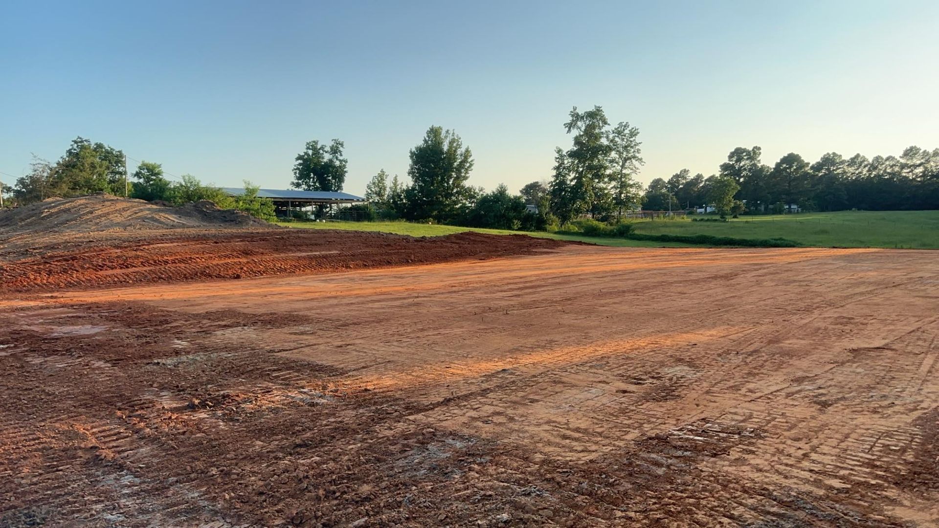 Brown dirt construction site with trees and a small building in the background.