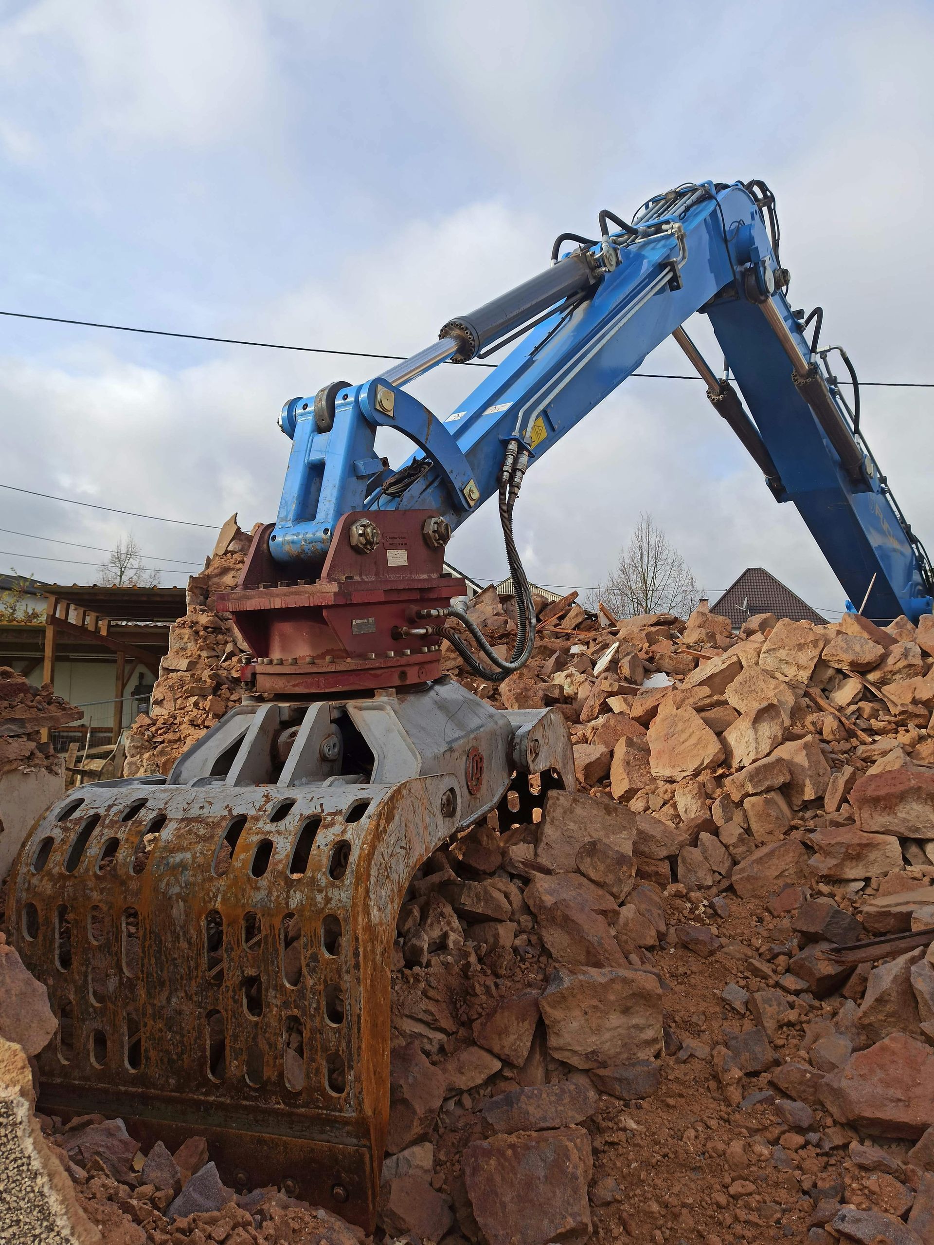 Blue excavator with demolition attachment, tearing apart brick rubble.