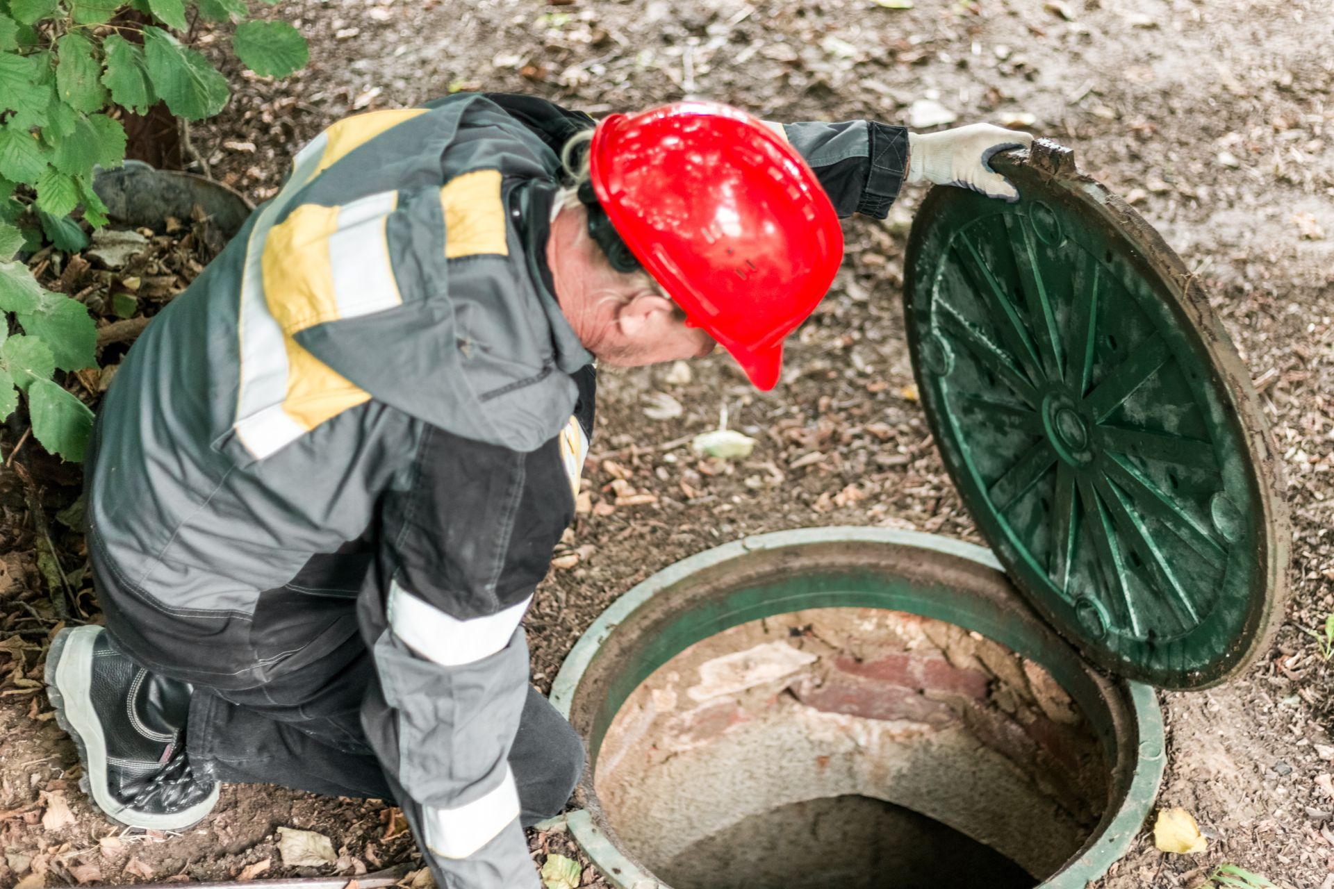 A man in a red hard hat leans over and inspects the underground shaft of a septic tank.