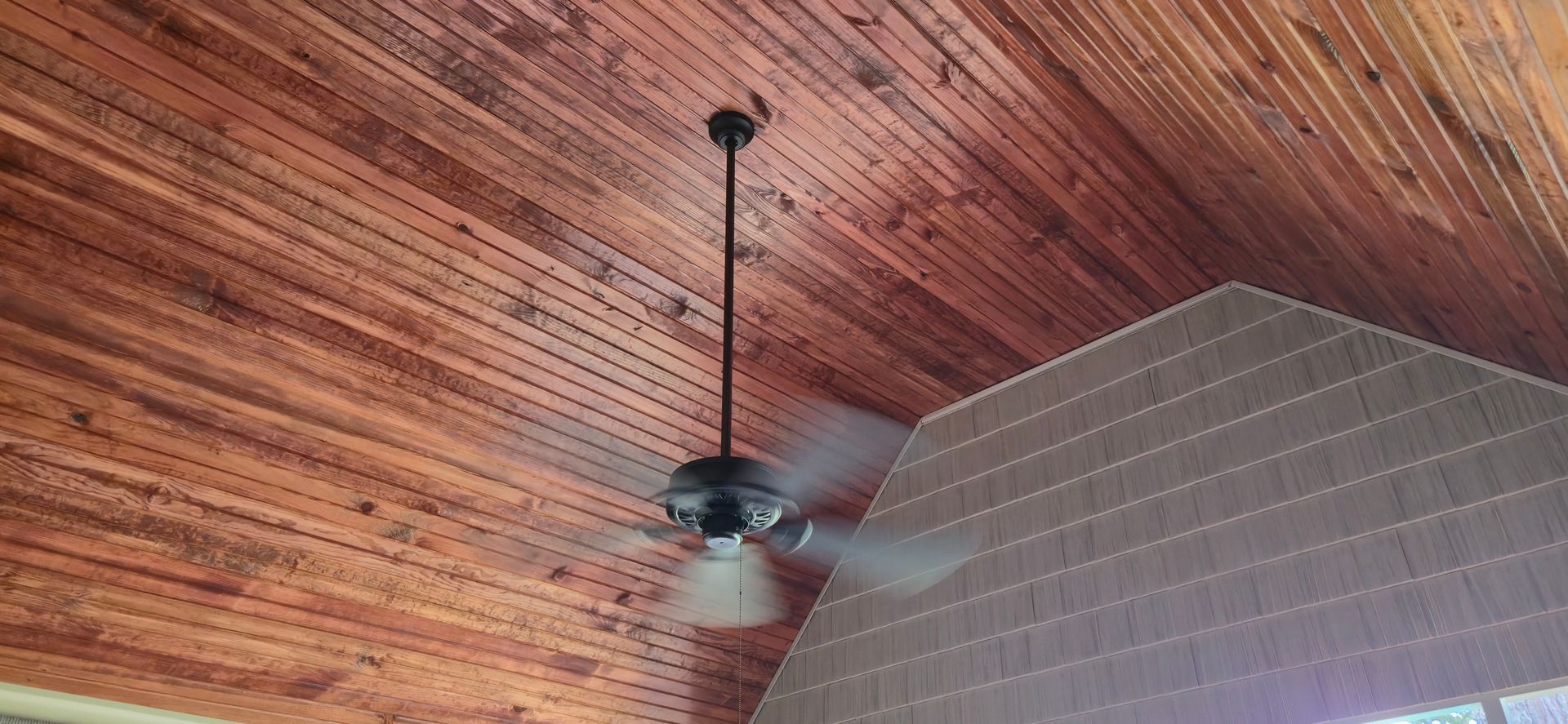 Ceiling fan with blurred blades against a wood plank ceiling. A light-colored wall section is also present.