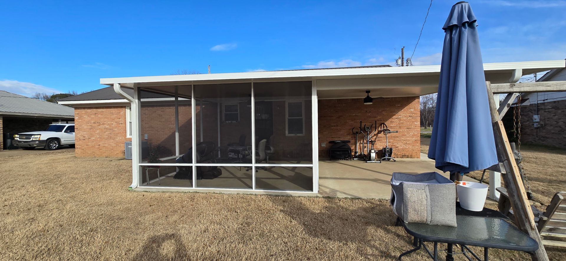 A screened-in porch next to a brick building under a clear blue sky. An umbrella stands nearby.