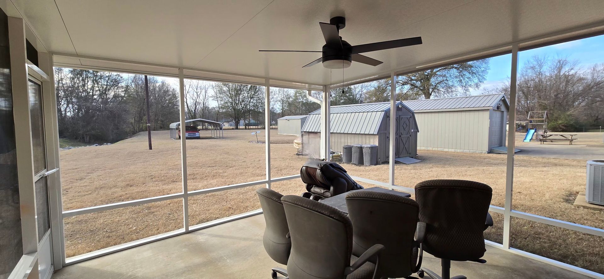 View from a screened porch towards a yard with sheds and a ceiling fan.