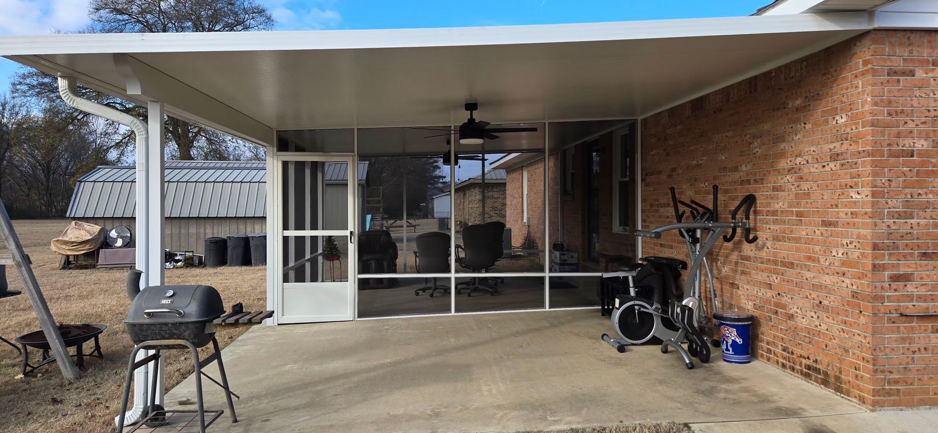 A covered patio with a grill, exercise equipment, and a screened enclosure. Brick wall on right.