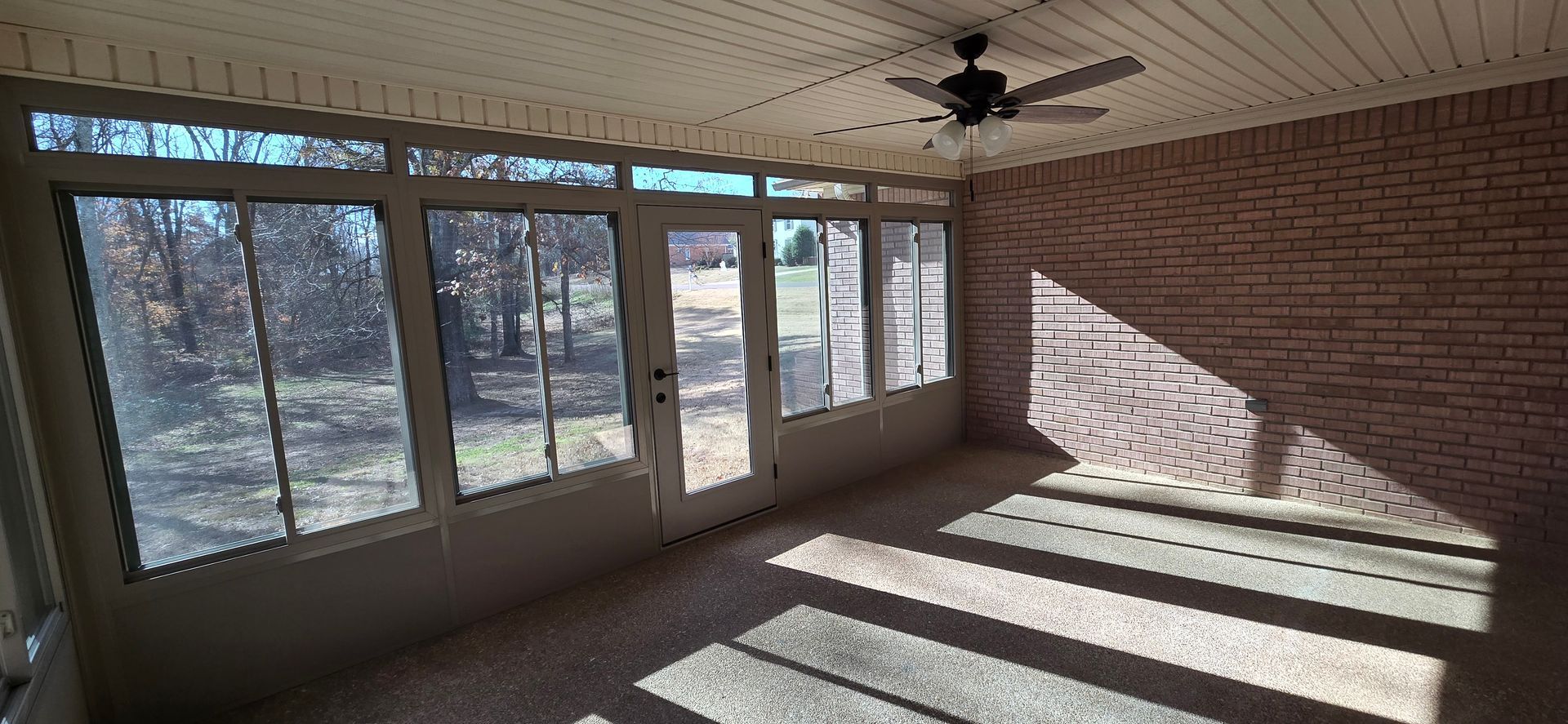 Sunroom with large windows, brick wall, and ceiling fan. Sunlight creates shadows on the floor.