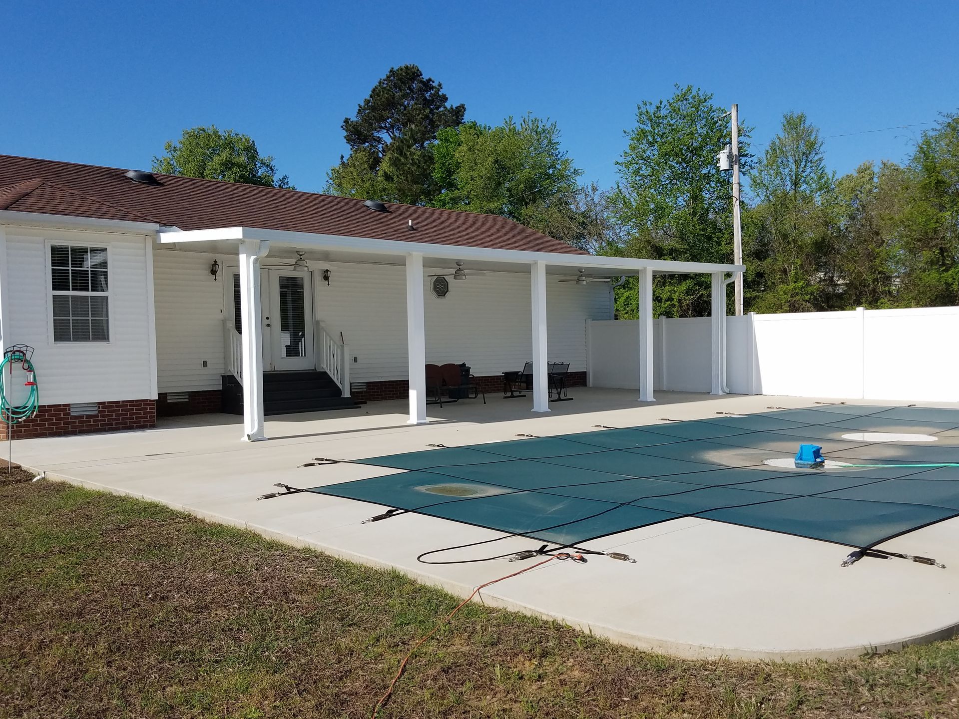 Poolside patio attached to a white house with a covered area, blue pool cover, and green lawn.