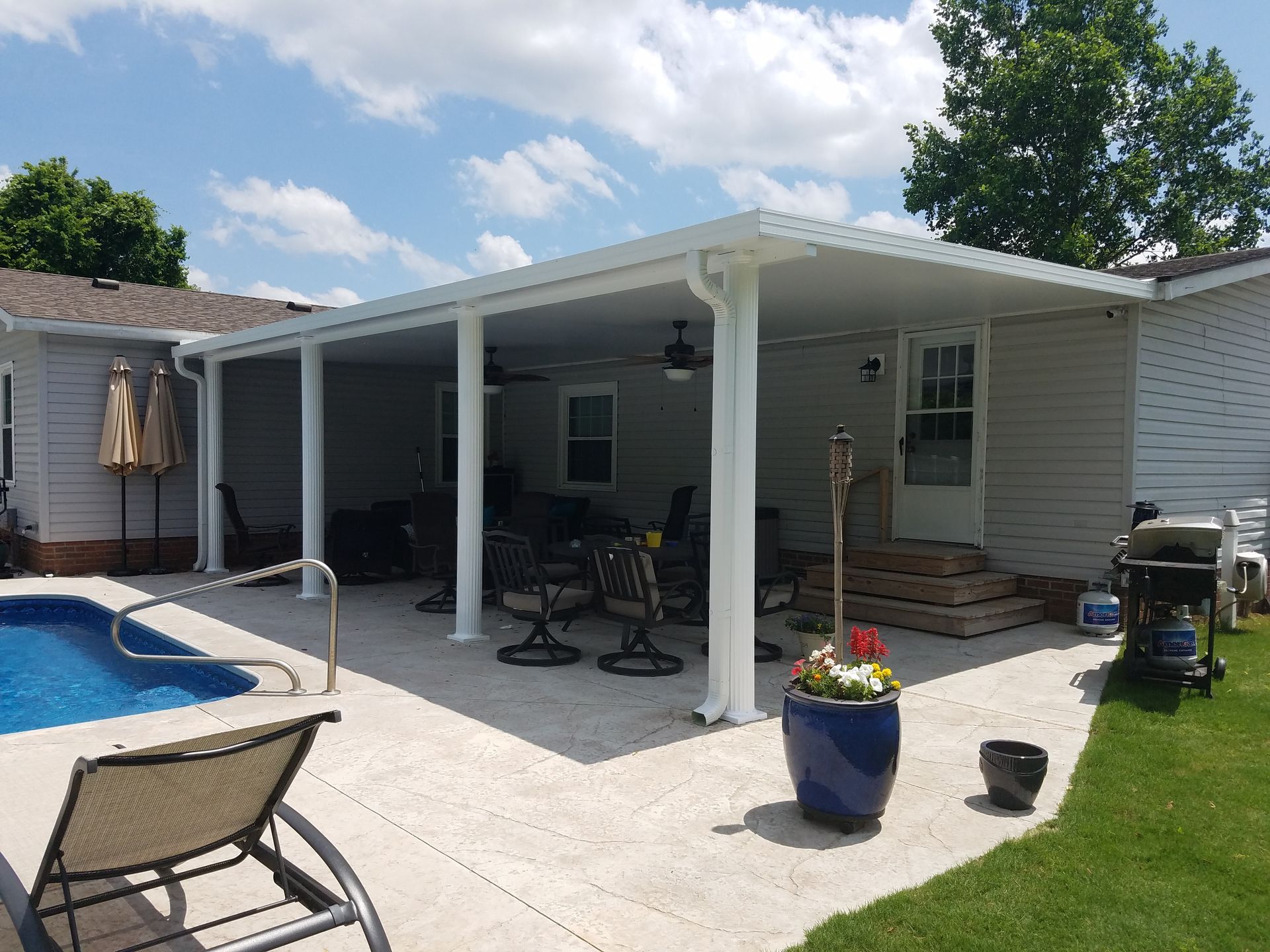Covered patio next to a pool and house with white columns, concrete floor, chairs, and barbecue grill.