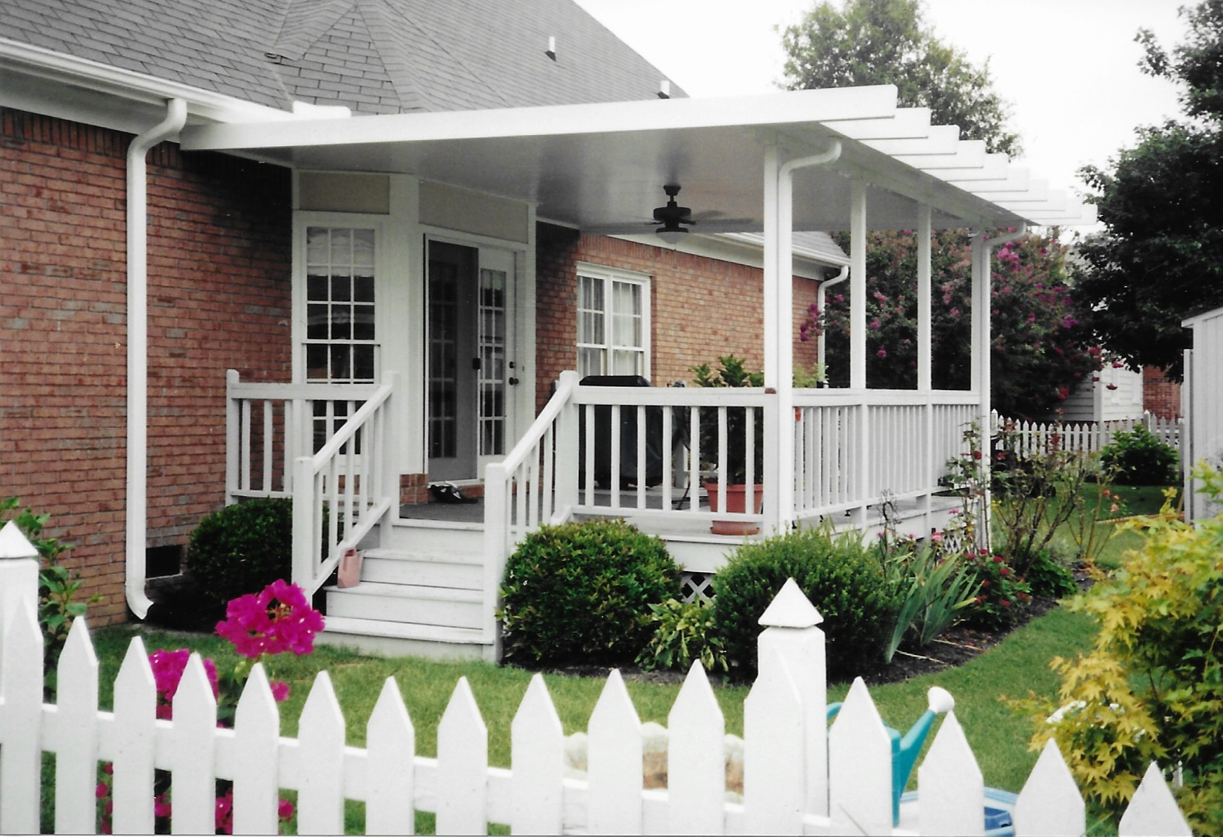Covered porch with white railings, steps, and a white picket fence in front of a brick house.