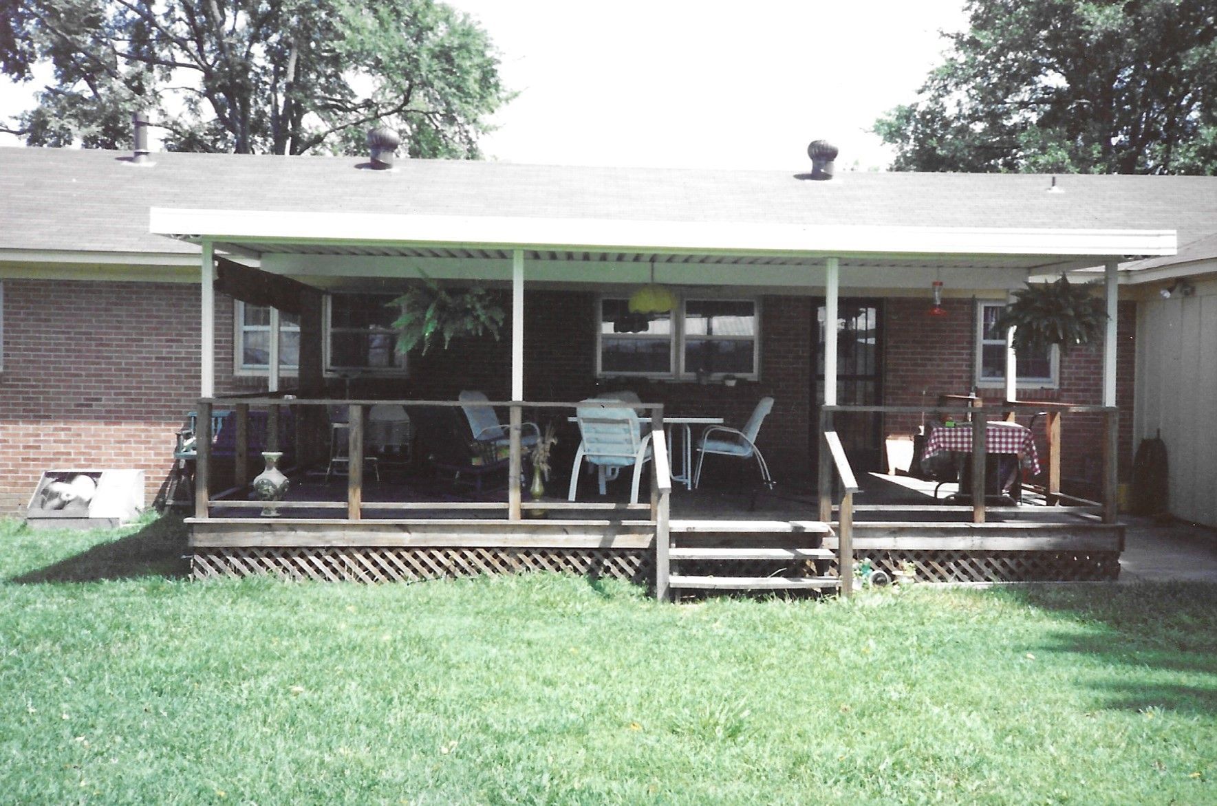 Covered wooden deck with chairs, table, and plants, attached to a brick house. Green lawn in front.