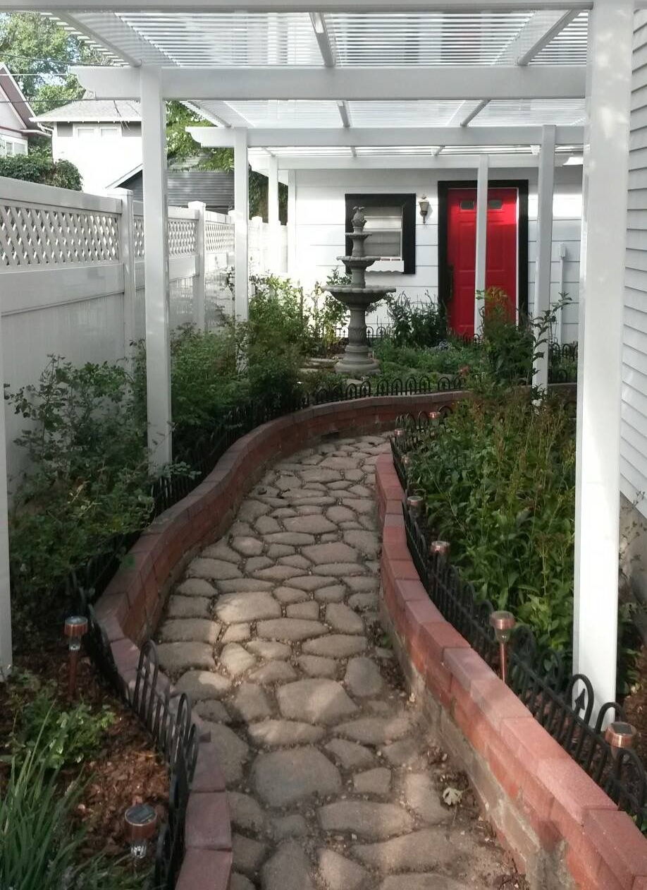 A cobblestone path winds through a garden, leading to a white building with a red door, under a white pergola.